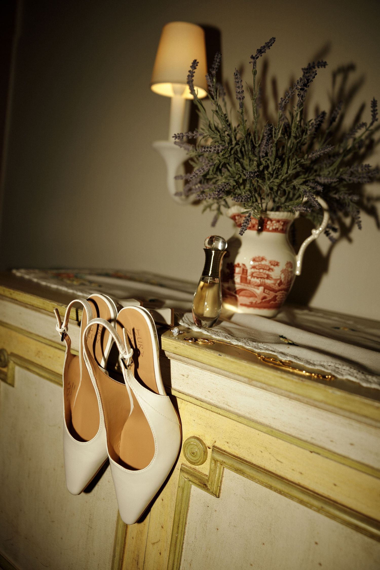 White bridal shoes displayed on vintage furniture with lavender bouquet and lamp in soft lighting.