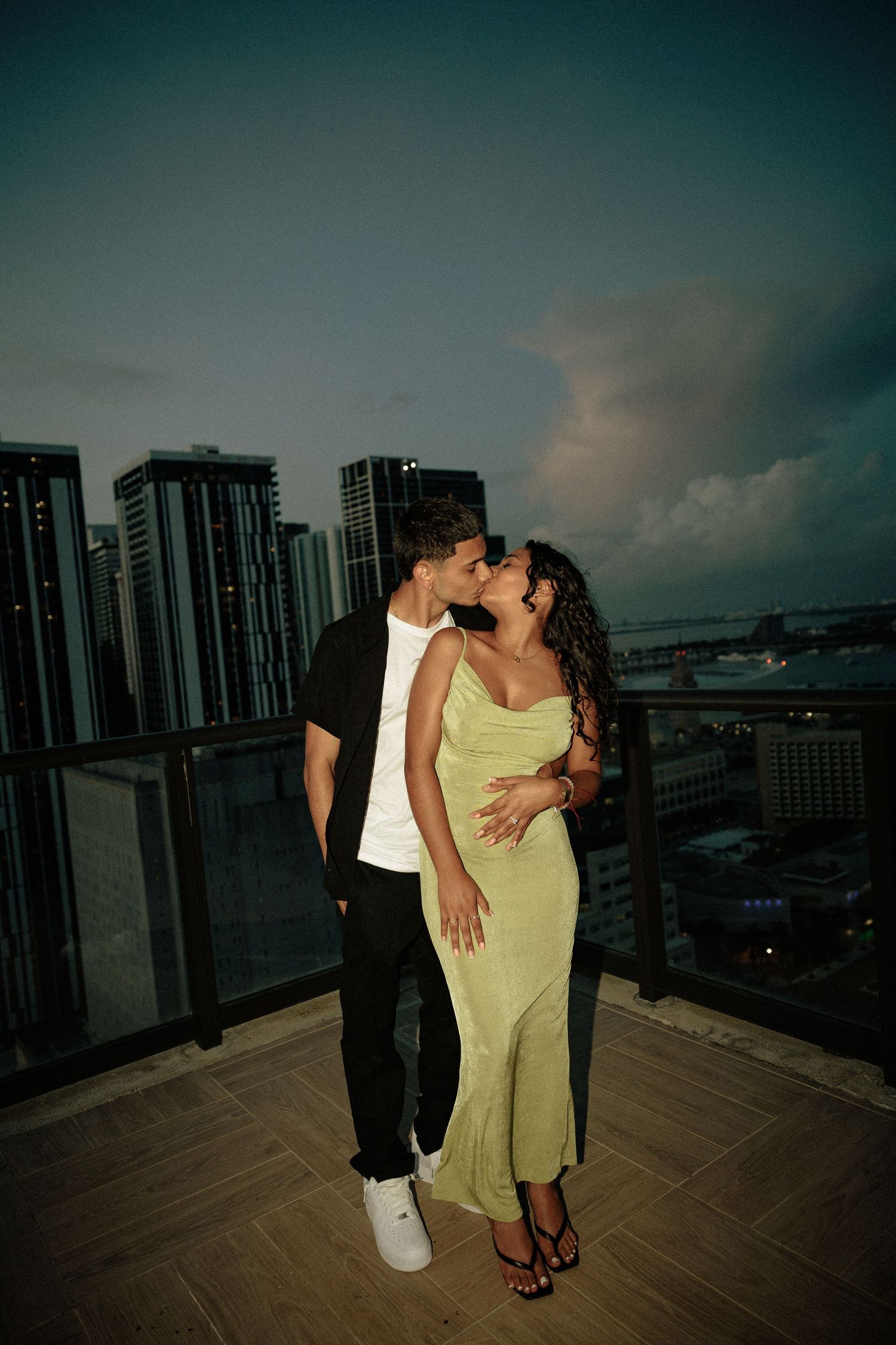 A romantic couple embraces on a rooftop balcony at sunset with city high-rises in the background.