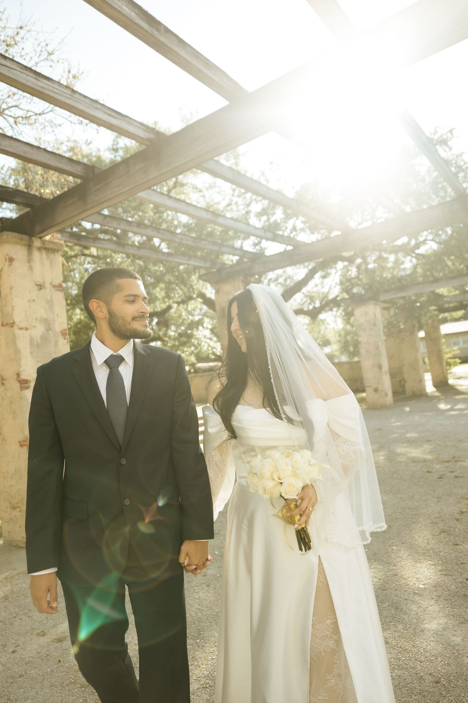 A couple in wedding attire walks outdoors under a sunlit pergola with stone pillars on a bright afternoon.