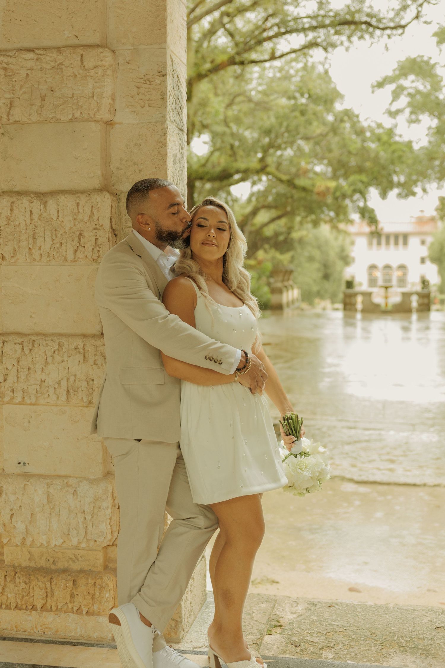 A romantic couple in white and beige outfits embrace under a stone archway during a warm summer photoshoot.