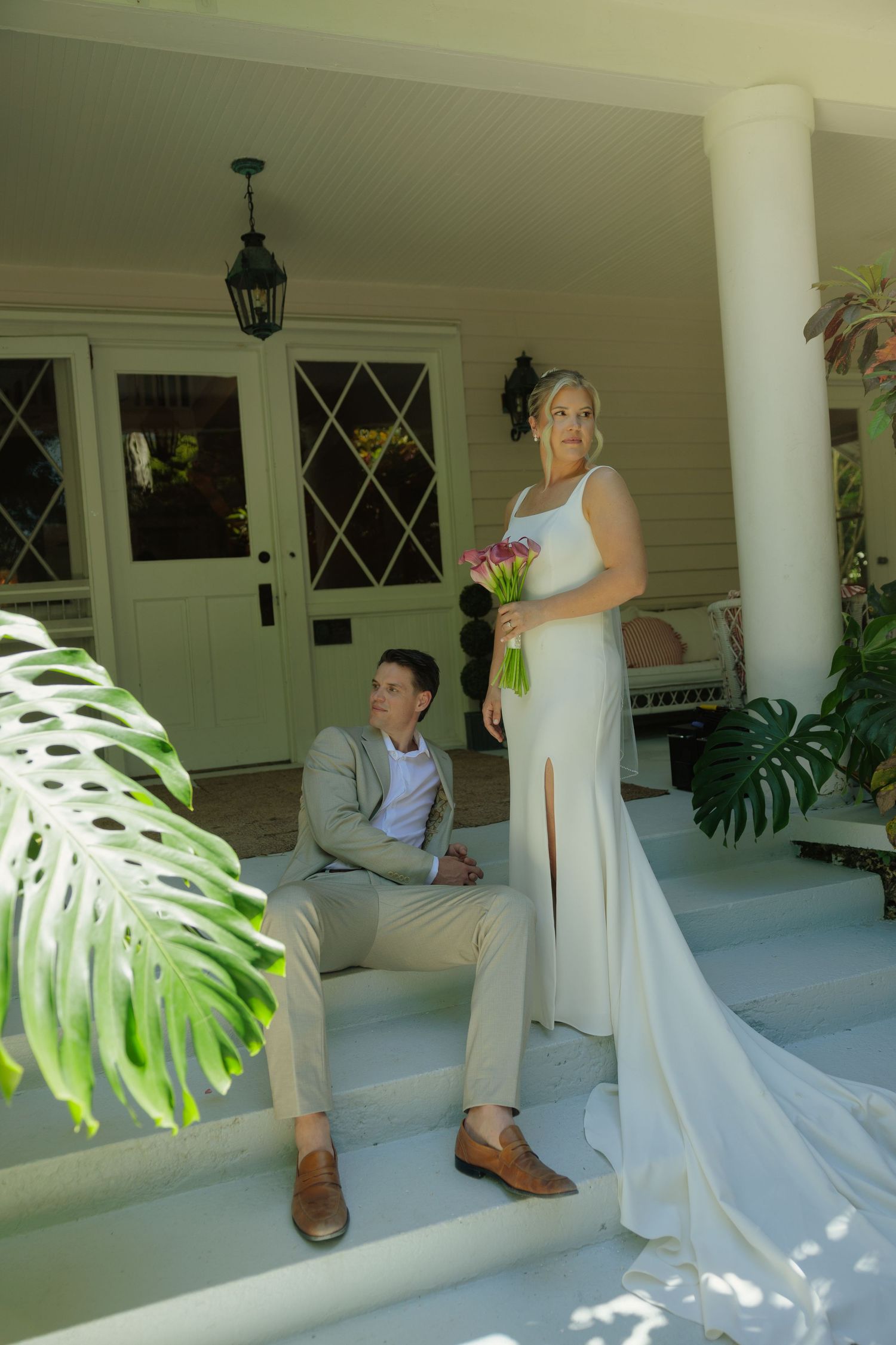 Bride in white gown holding pink bouquet stands on porch steps beside seated groom in tan suit at elegant venue.