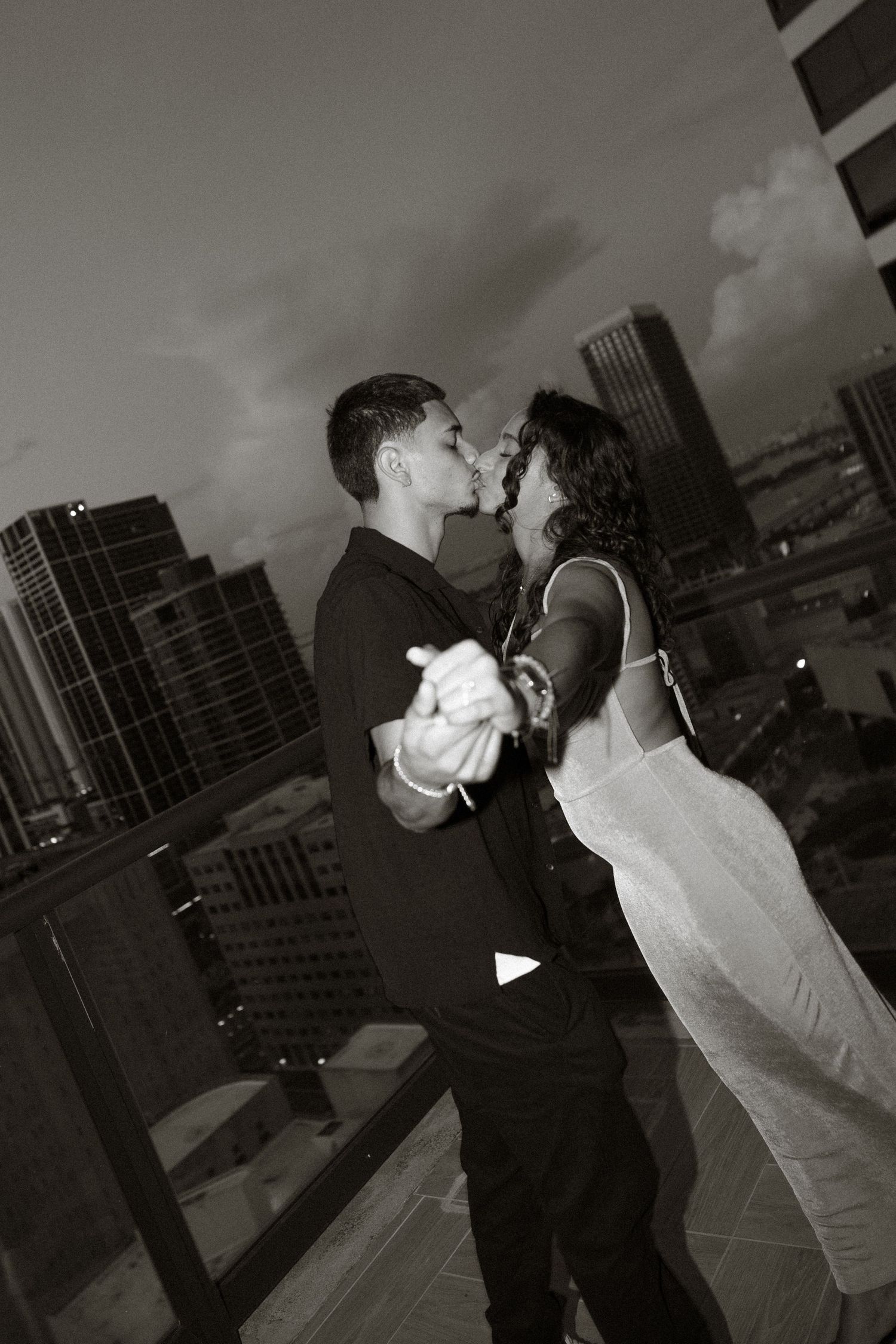 A couple shares a romantic kiss at sunset on a rooftop terrace with city buildings in the background.