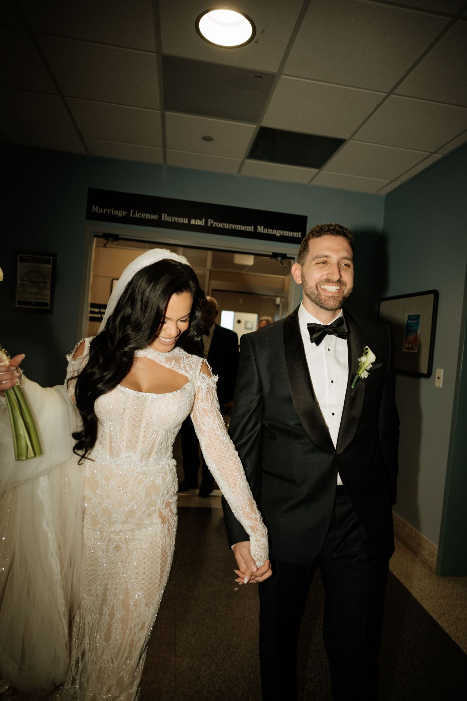 A couple in formal wedding attire walks down a hallway holding hands, with the bride wearing a sparkly white gown.