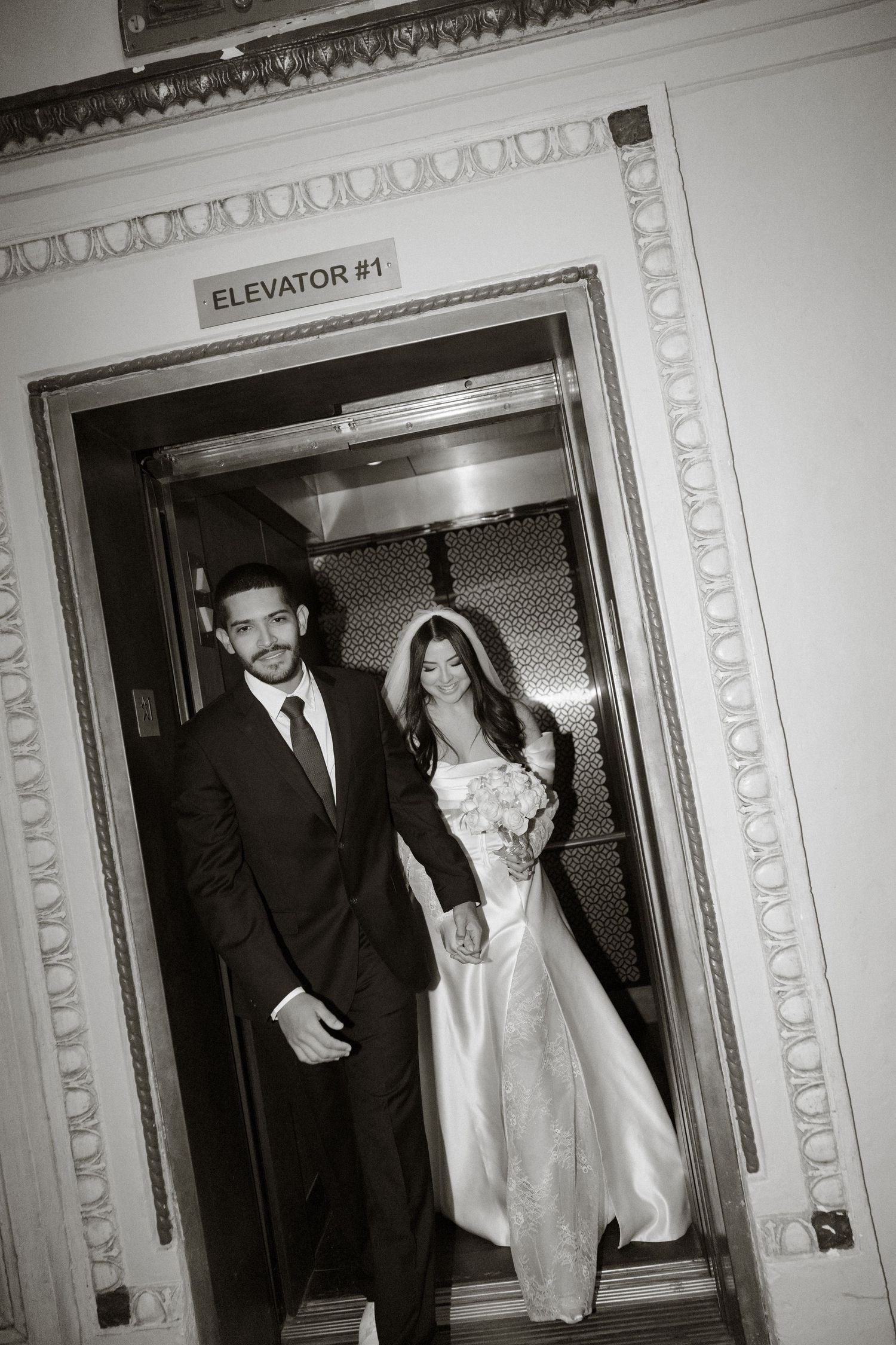 A bride and groom emerge from an ornate vintage elevator in black and white wedding photography.
