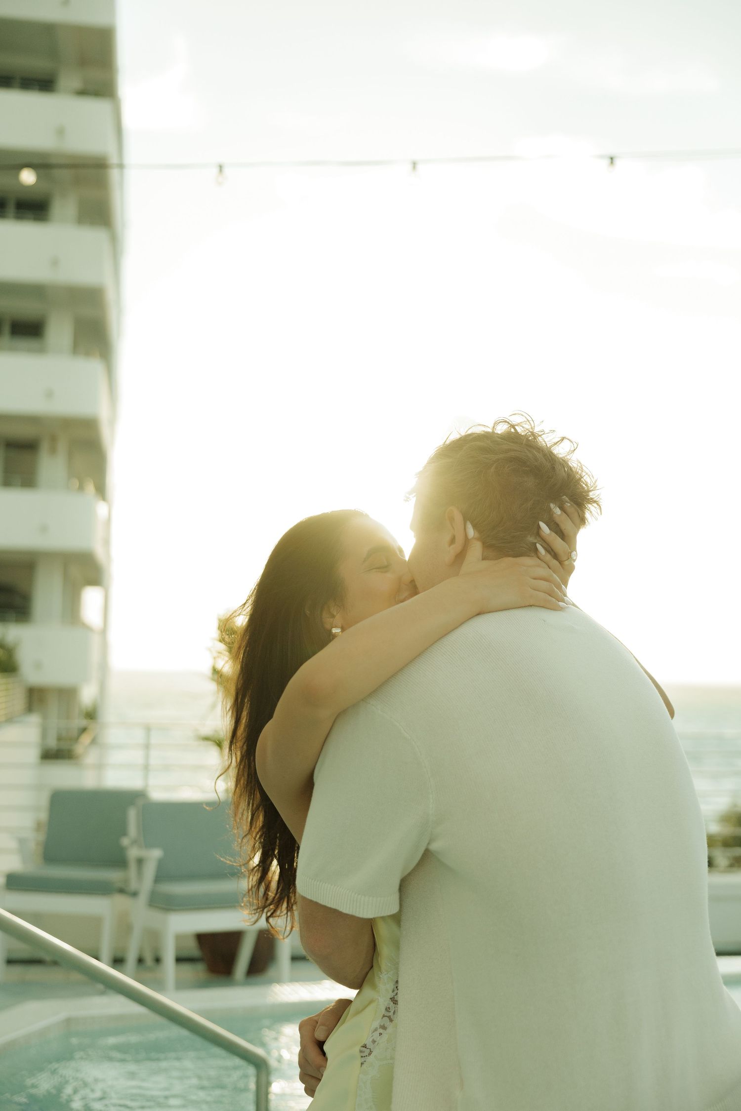 Couple sharing a romantic kiss on a sunlit hotel balcony overlooking the ocean at golden hour.