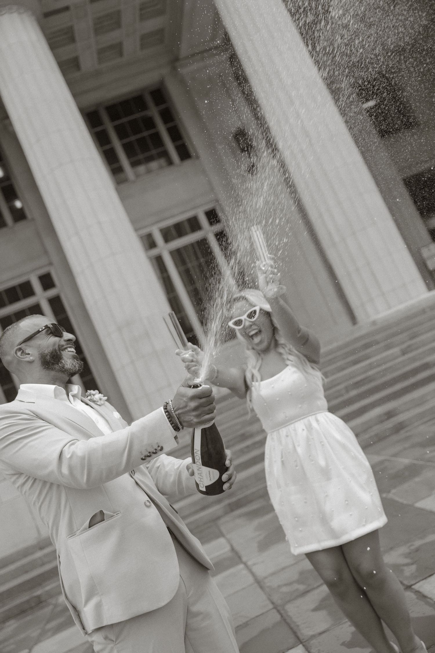 Black and white photo of a couple in white attire celebrating with champagne spray on building steps.