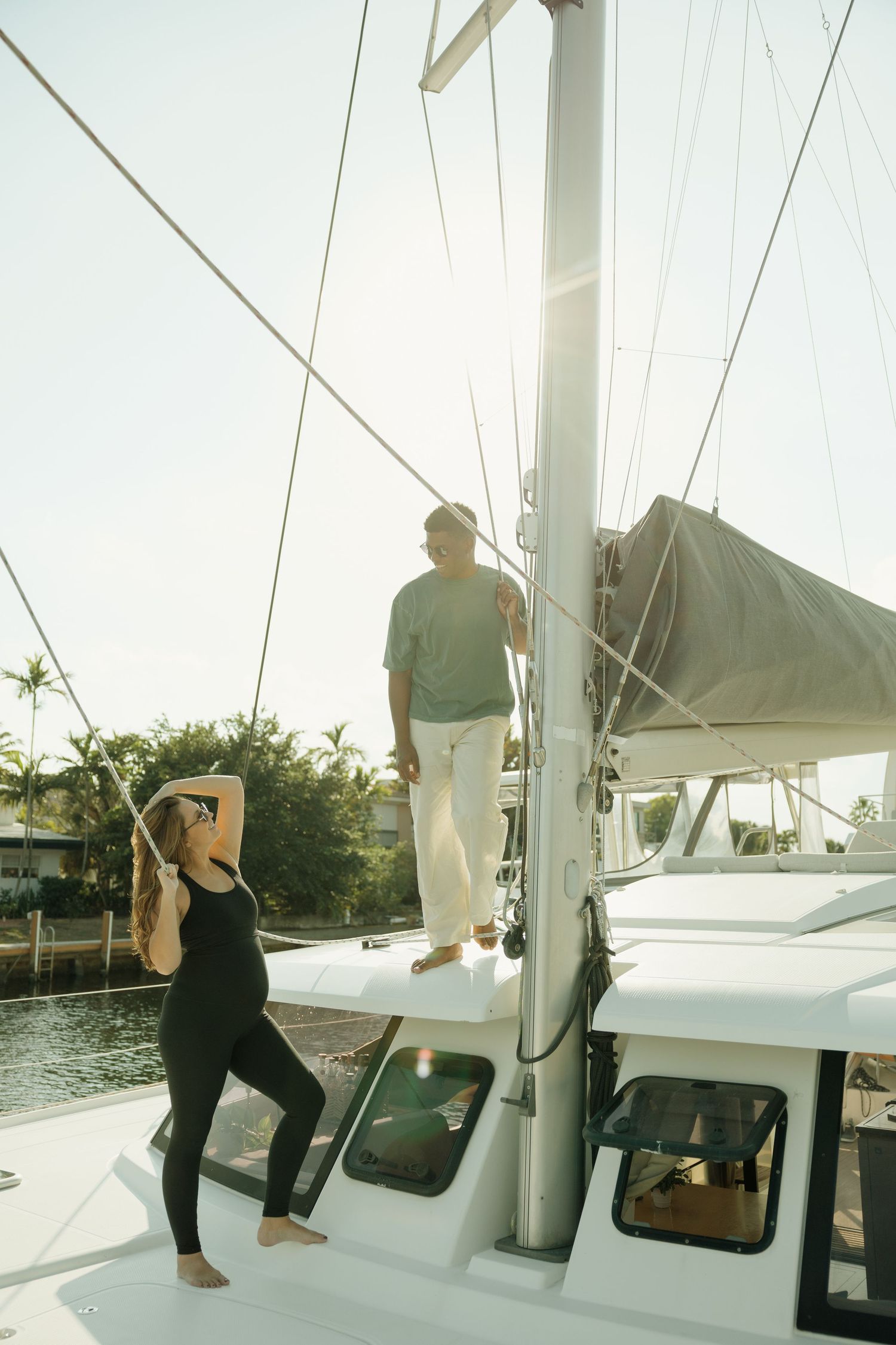 Couple relaxing on a white sailboat near a tropical marina, woman stretching in black outfit, man standing by the mast.