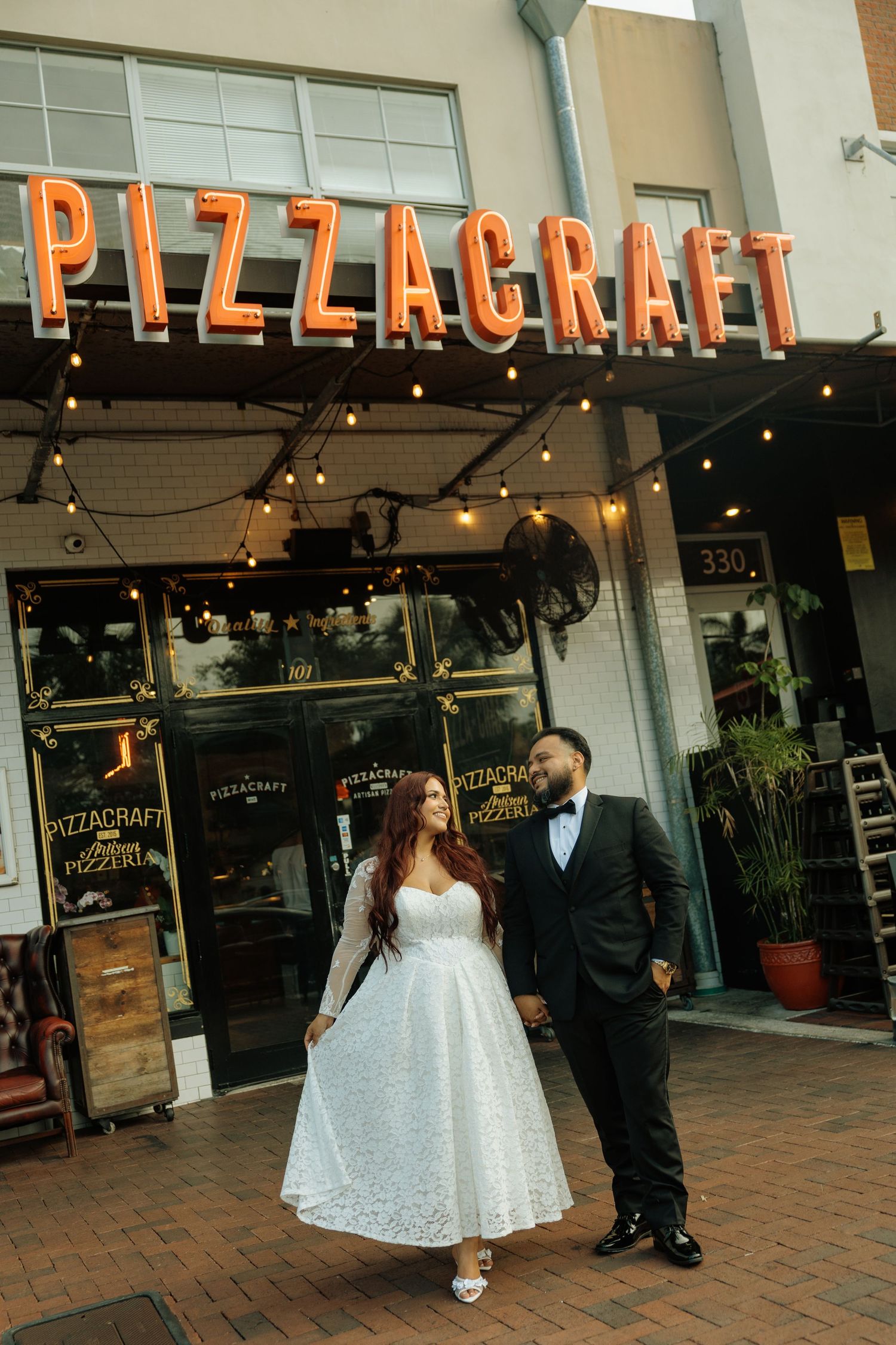 Bride and groom pose romantically outside Pizzacraft restaurant with glowing sign and string lights.