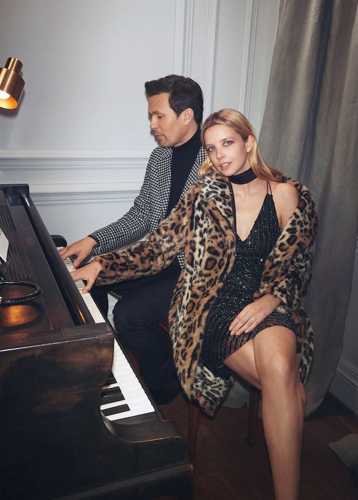A glamorous couple poses at a grand piano, woman in leopard coat and sequin dress, man in houndstooth blazer.