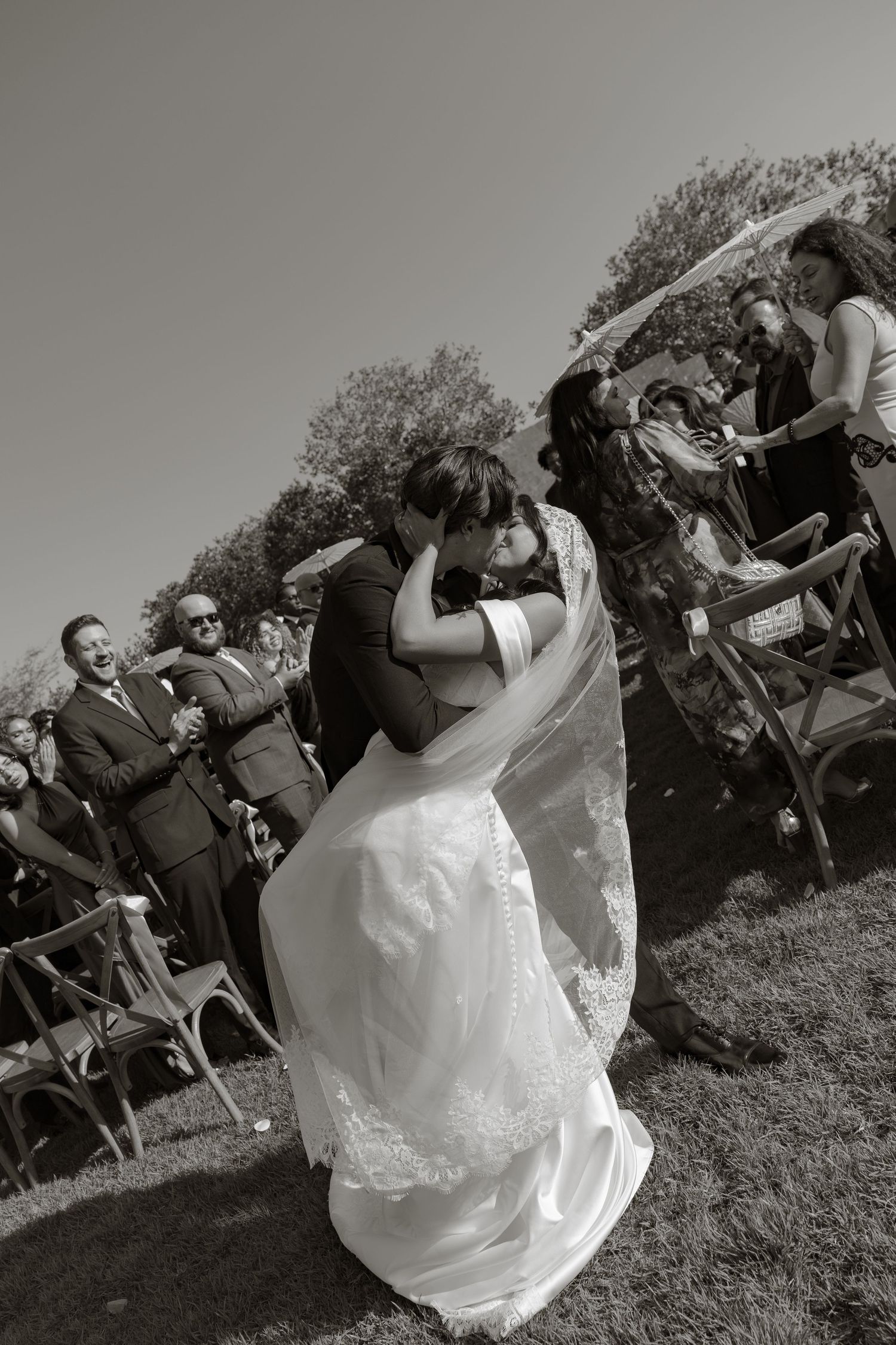 Black and white photo of a bride and groom sharing their first kiss at an outdoor wedding ceremony with guests cheering.