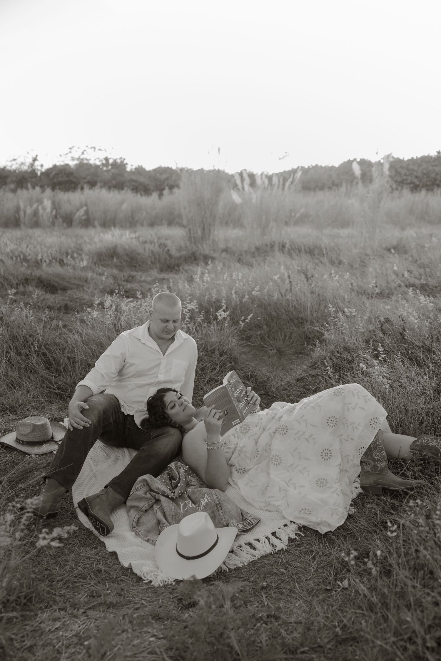 Black and white photo of a couple relaxing in a field, woman lying down reading a book while man sits beside her.