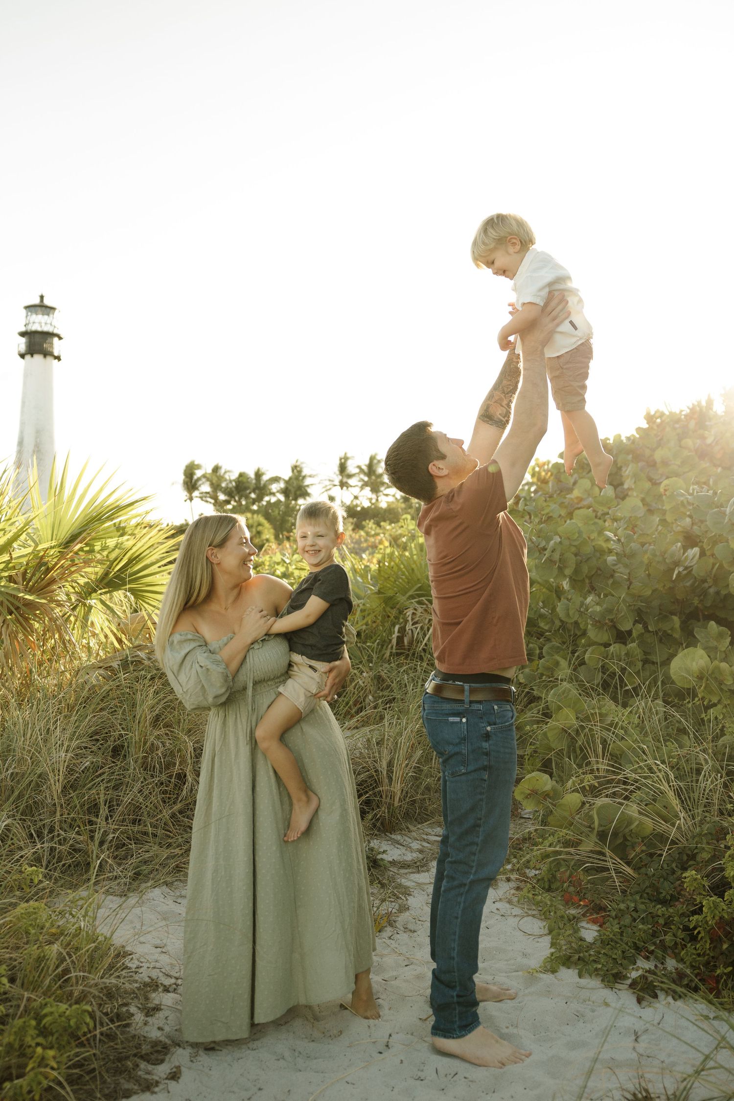 A family enjoys a sunset moment together on a beach path with a lighthouse in the background.