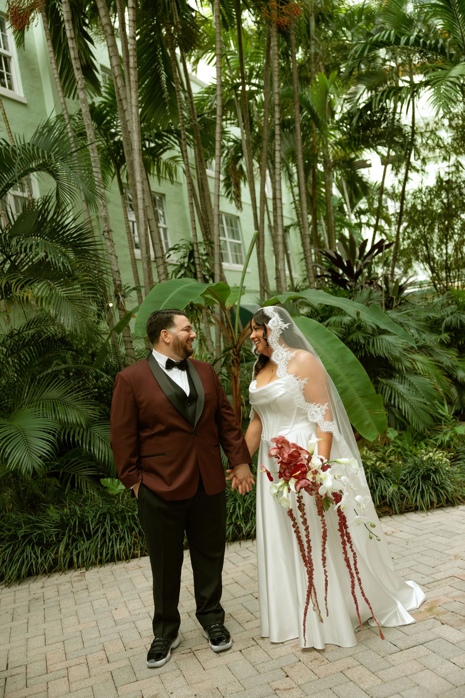 A wedding couple shares romantic moments in a lush tropical garden setting with palm trees and brick pathways.