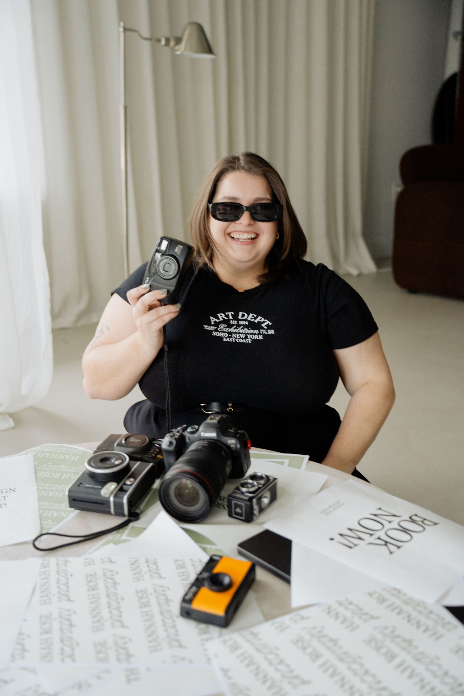 Photographer sits at desk with vintage and modern cameras, sunglasses, and papers spread out on white surface.