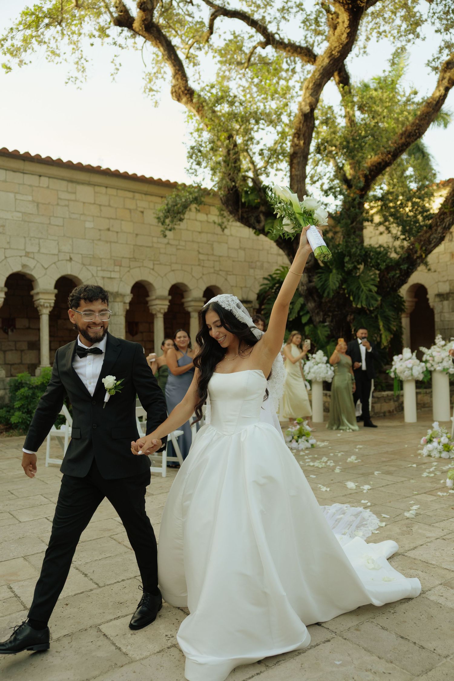 Bride raises bouquet triumphantly as groom smiles, exiting outdoor wedding ceremony at stone venue.
