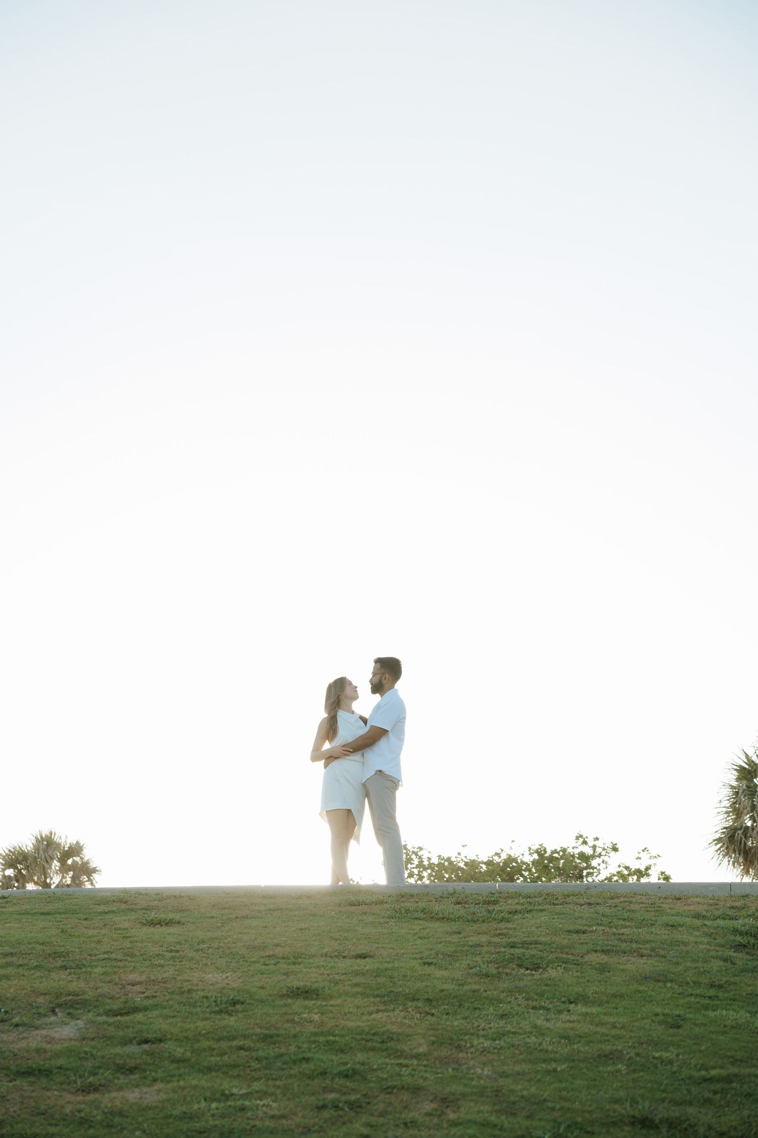 A couple embraces on a grassy hilltop silhouetted against a bright white sky at sunset.