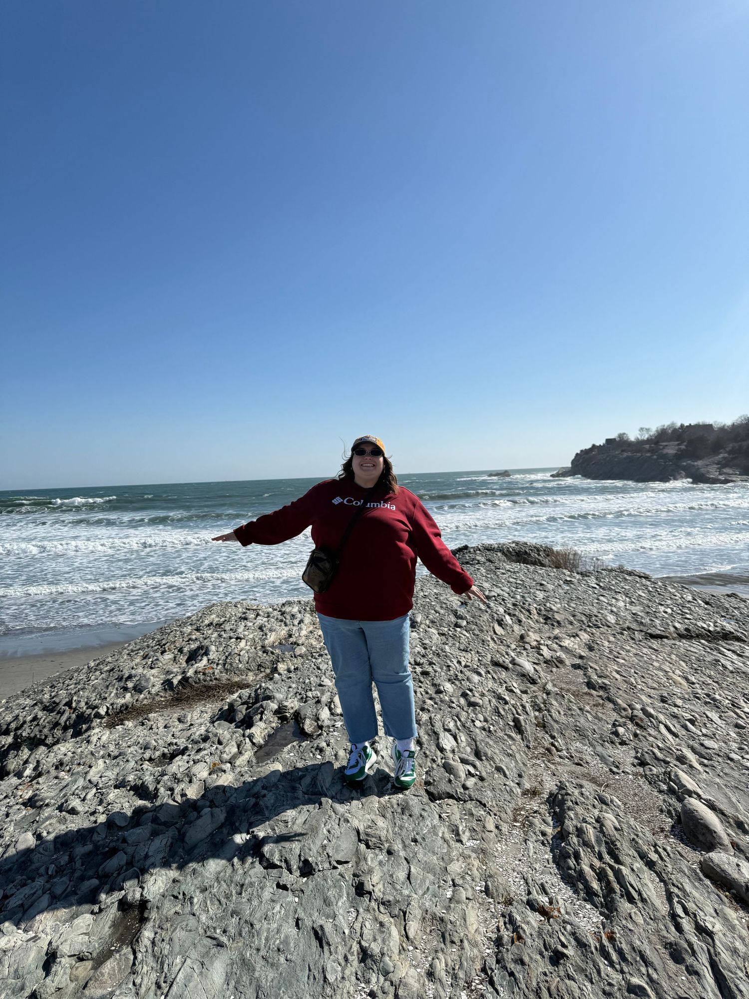 Someone in a red jacket and jeans stands on rocky coastal cliffs with crashing ocean waves and blue skies in the background.