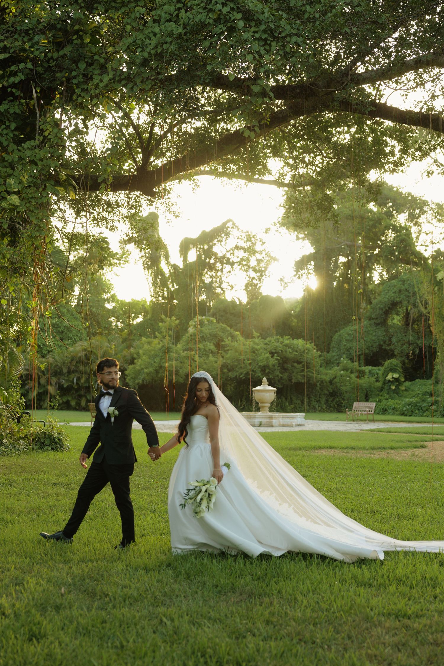 Bride and groom walk hand-in-hand on lush green lawn under large oak tree at golden hour wedding.
