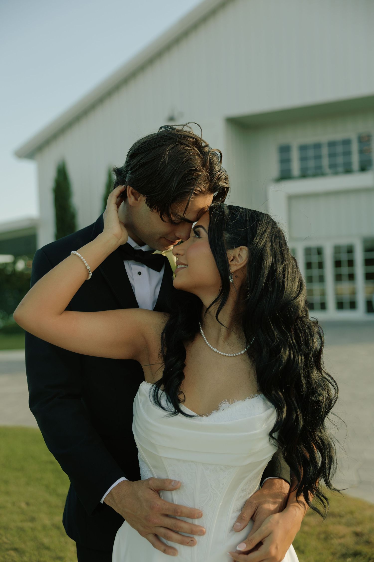A bride and groom share a romantic almost-kiss moment outdoors, dressed in a white gown and black tuxedo.