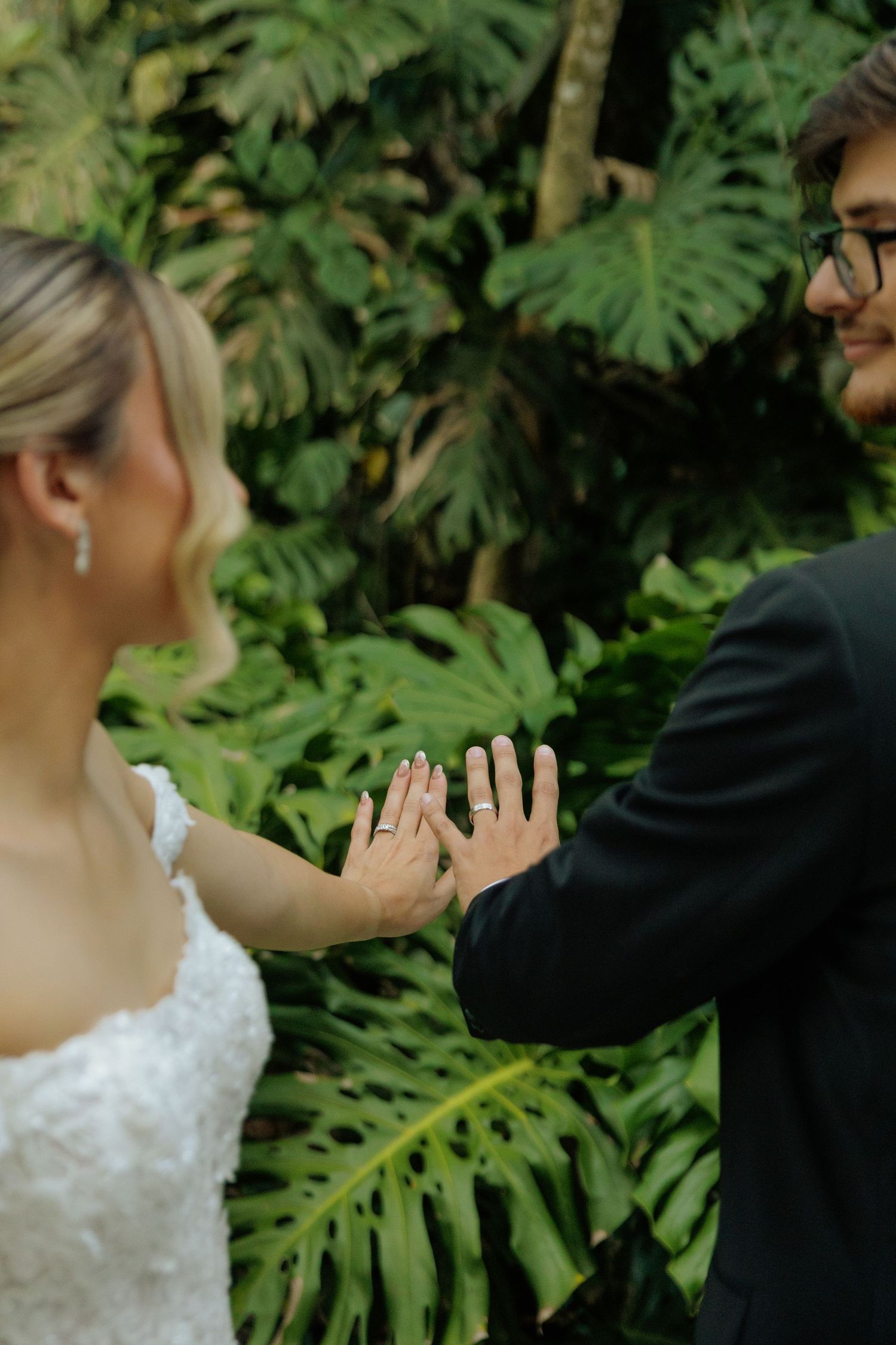 Bride and groom comparing hands with wedding rings against lush tropical green foliage backdrop.