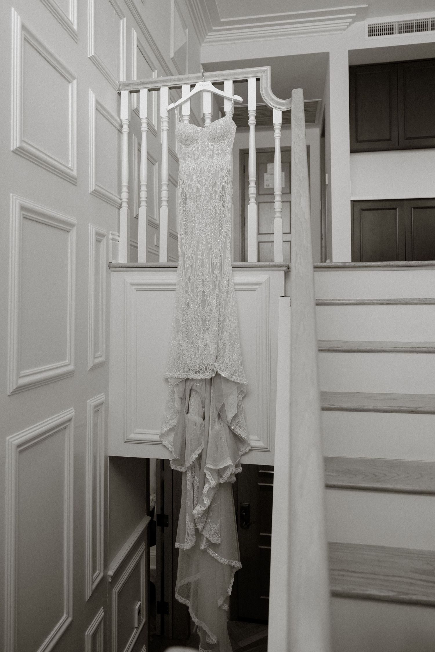 A white lace wedding dress hanging on a hanger from a staircase banister in an elegant home interior.