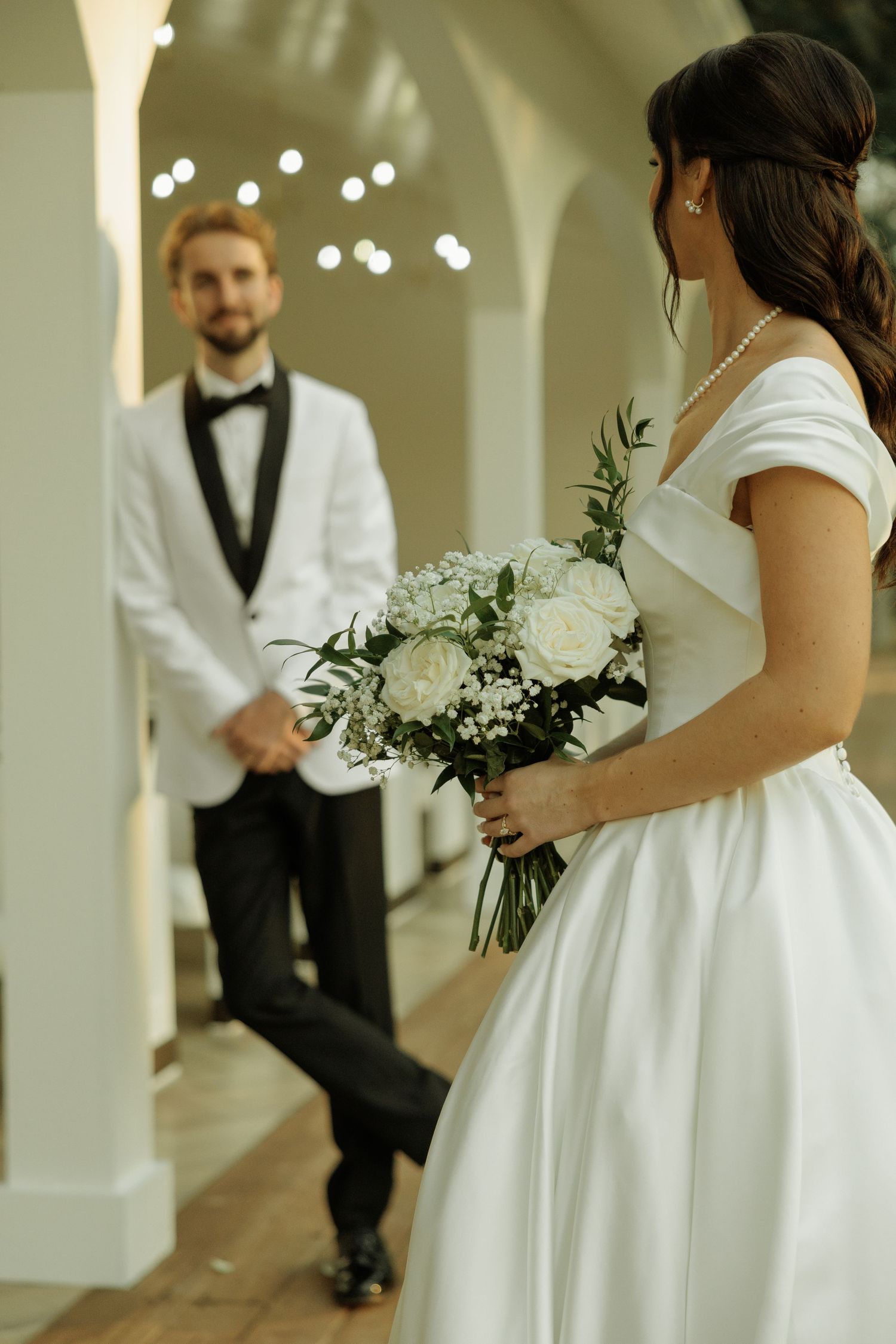 A romantic first look moment between a bride in white dress and groom in black vest at an indoor venue.