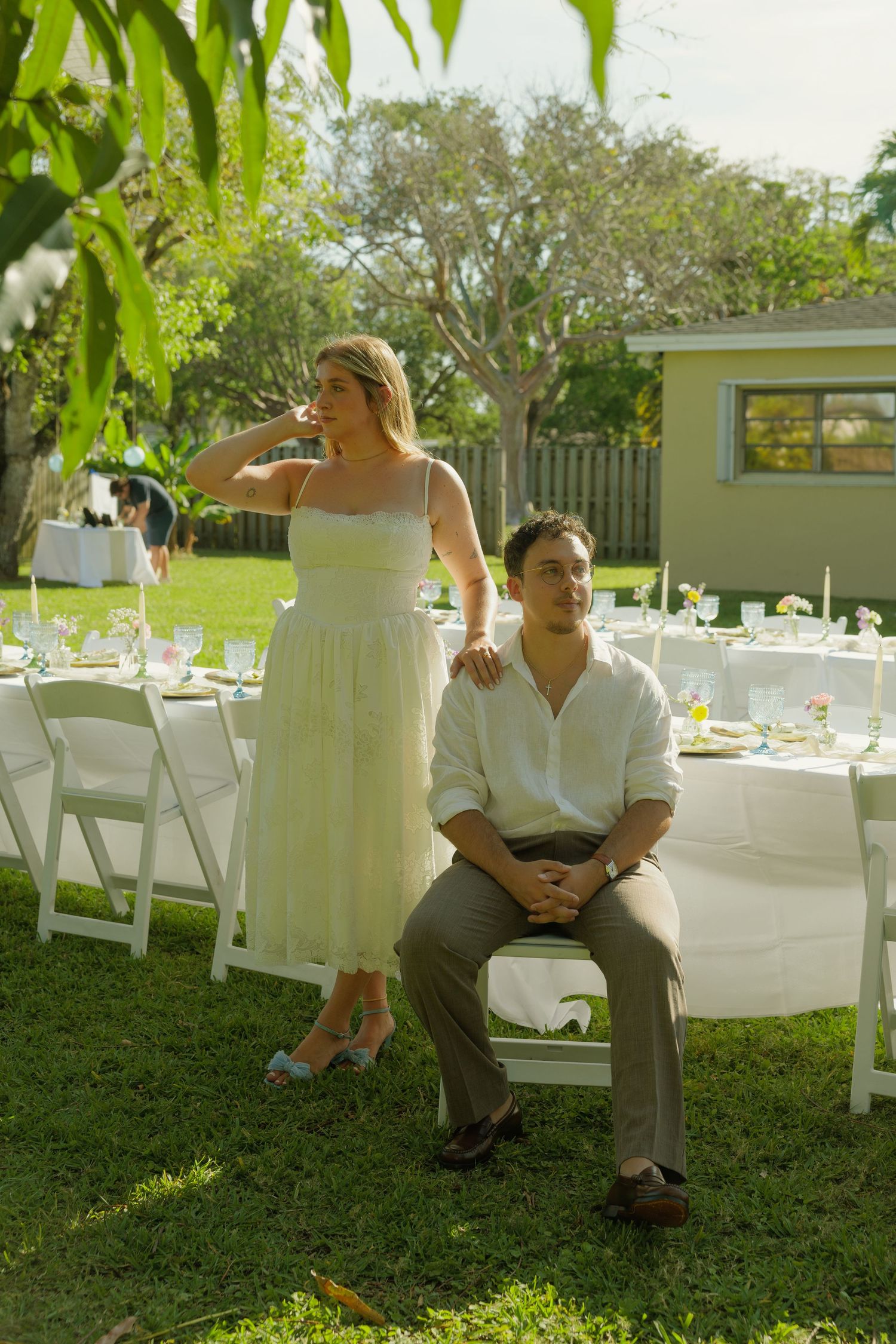 Couple in white attire posing at an outdoor wedding reception with decorated tables on a sunny green lawn.