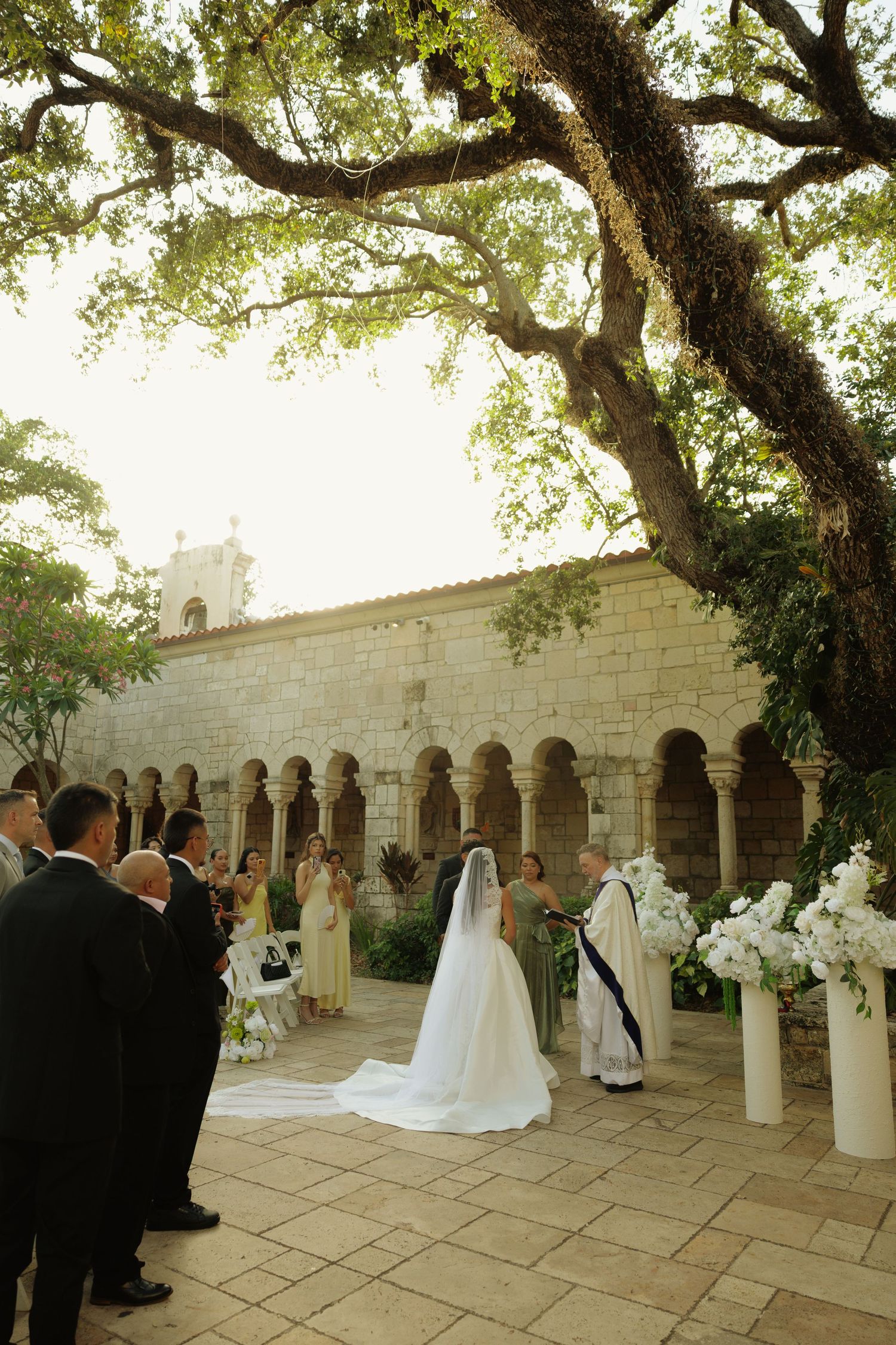 Outdoor wedding ceremony in a historic stone courtyard with arched columns, white floral arrangements, and a bride in a long veil.