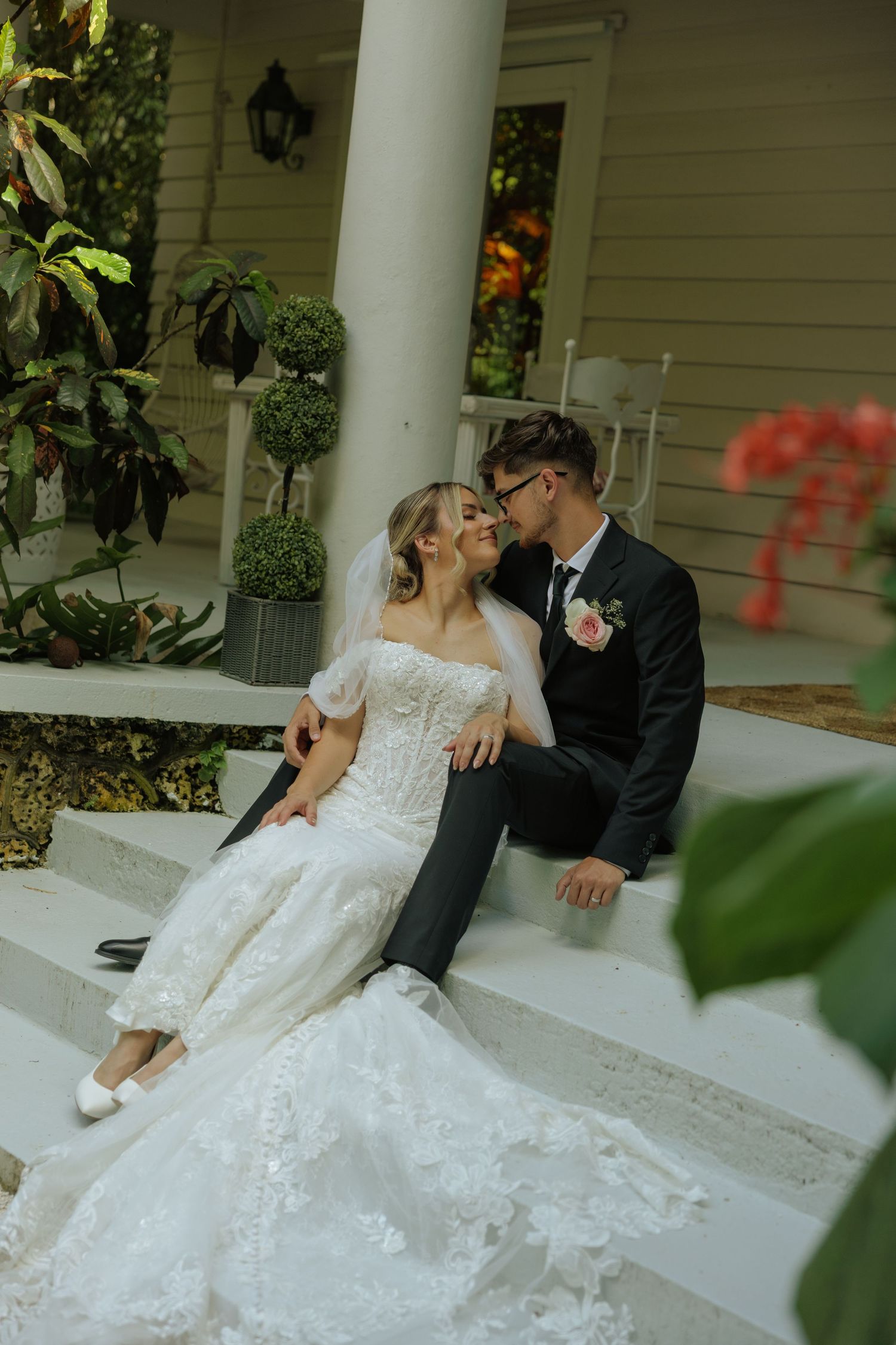 Bride and groom sit together on white porch steps, sharing a romantic moment in wedding attire.