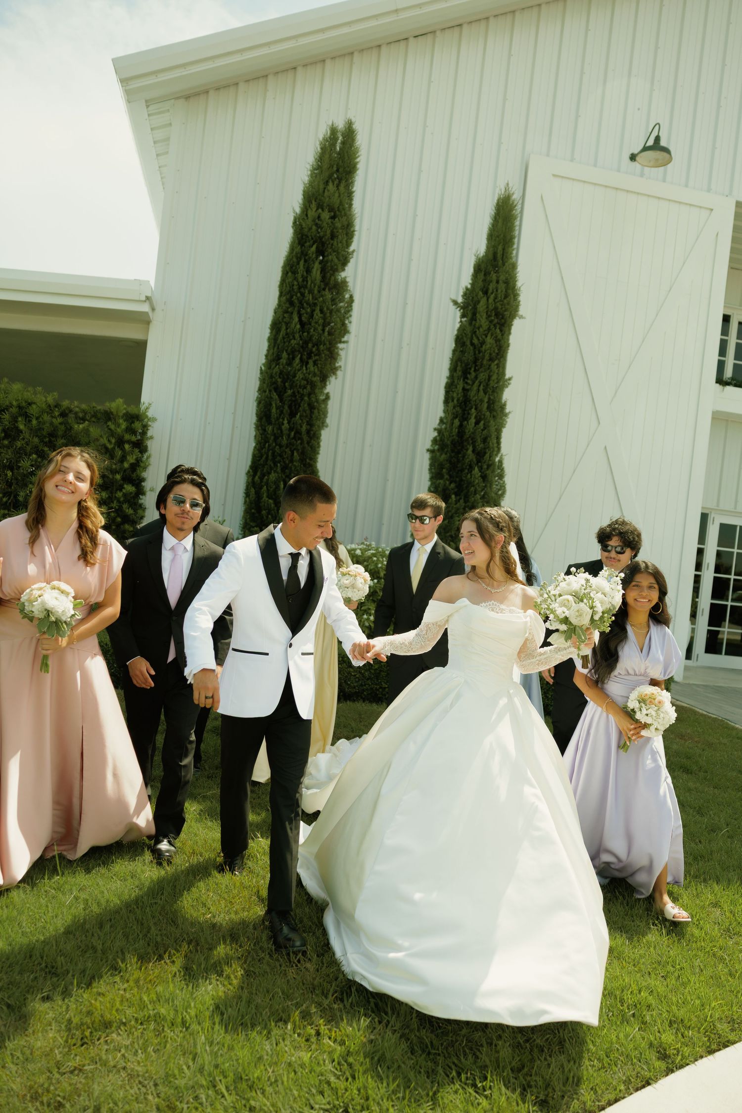 Bride and groom hold hands walking with wedding party outside white barn venue on sunny day.