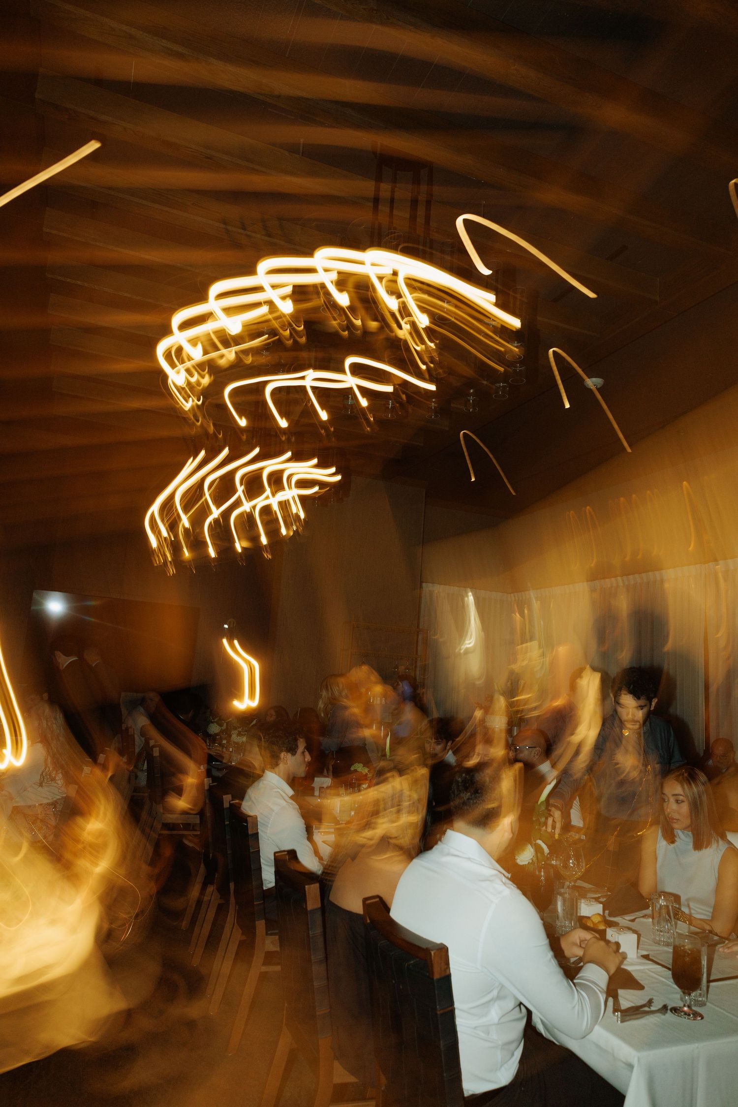 Blurred motion long exposure photo of diners in a warmly lit restaurant with glowing light streaks overhead.
