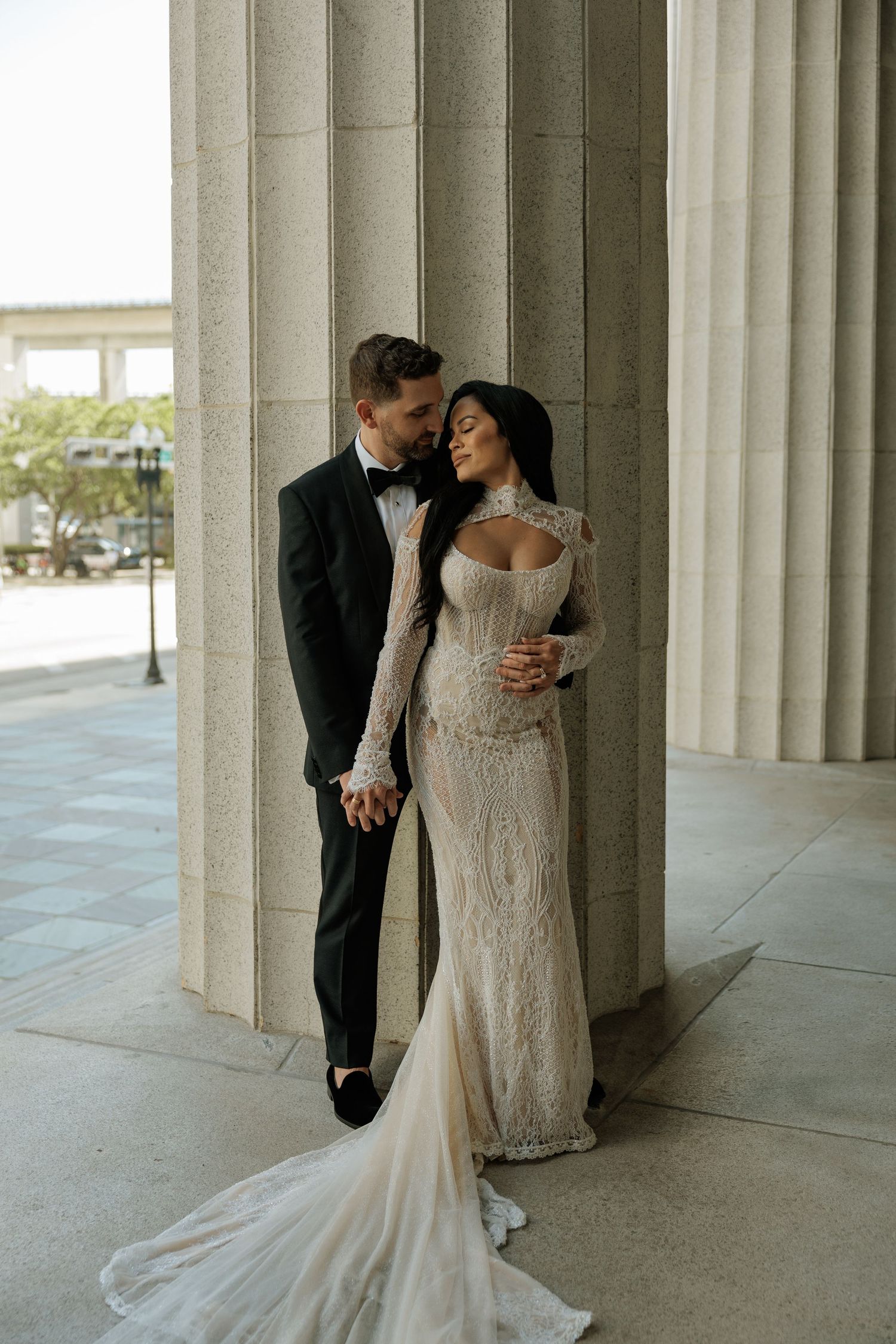 Elegant bride and groom pose between stone columns, she in lace gown with train, he in black tuxedo.