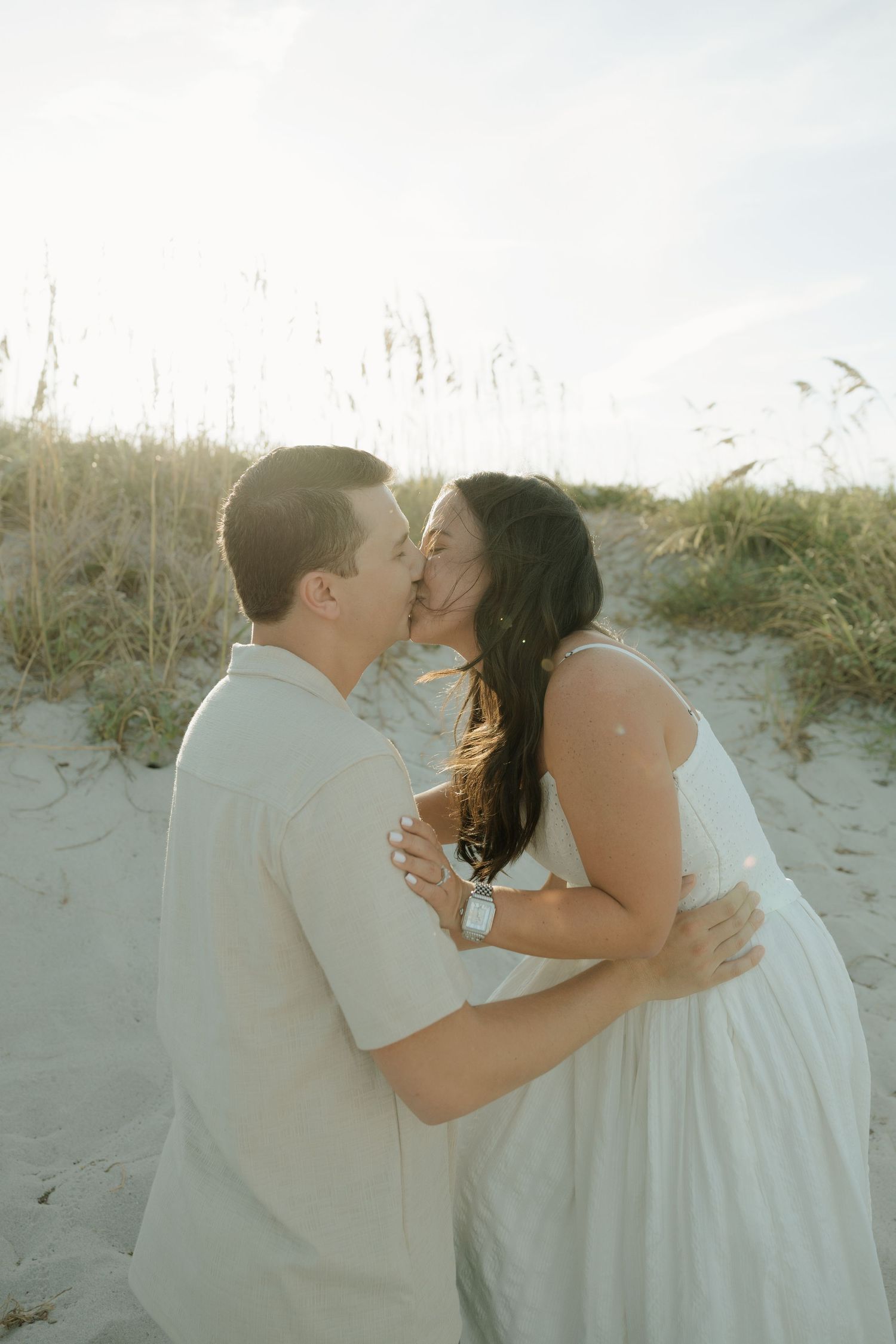 A romantic couple shares a tender kiss on a sandy beach with sea grass swaying in the background.