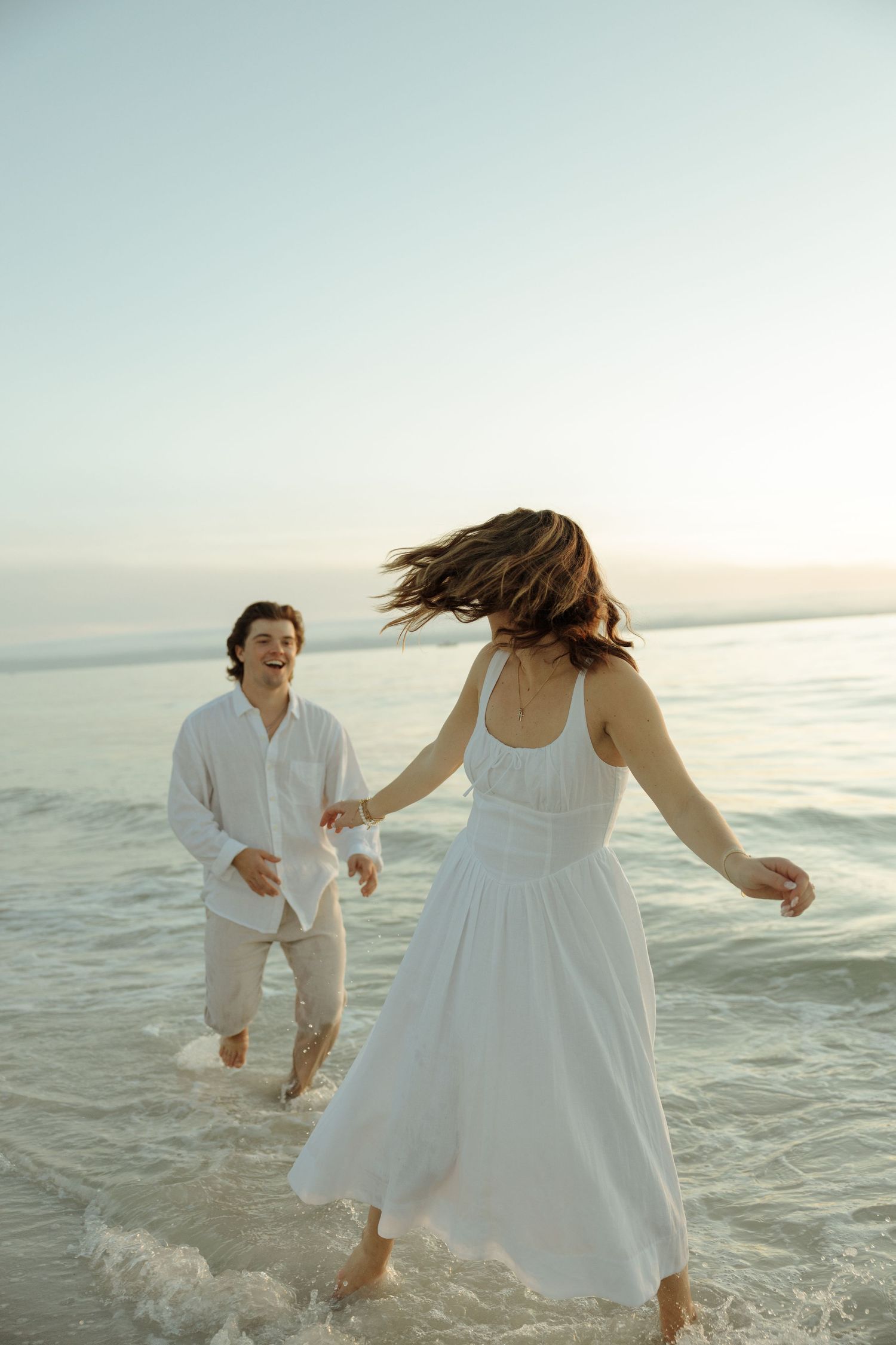 Playful couple in white attire splashing and dancing in shallow ocean waves at sunset.