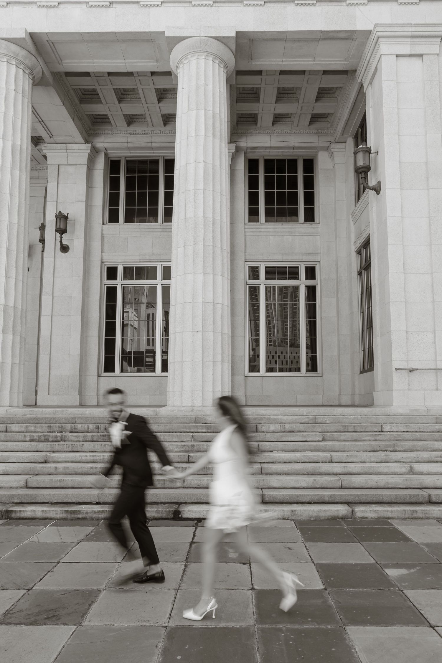 Black and white photo of a couple holding hands and walking past a grand neoclassical building with tall columns.