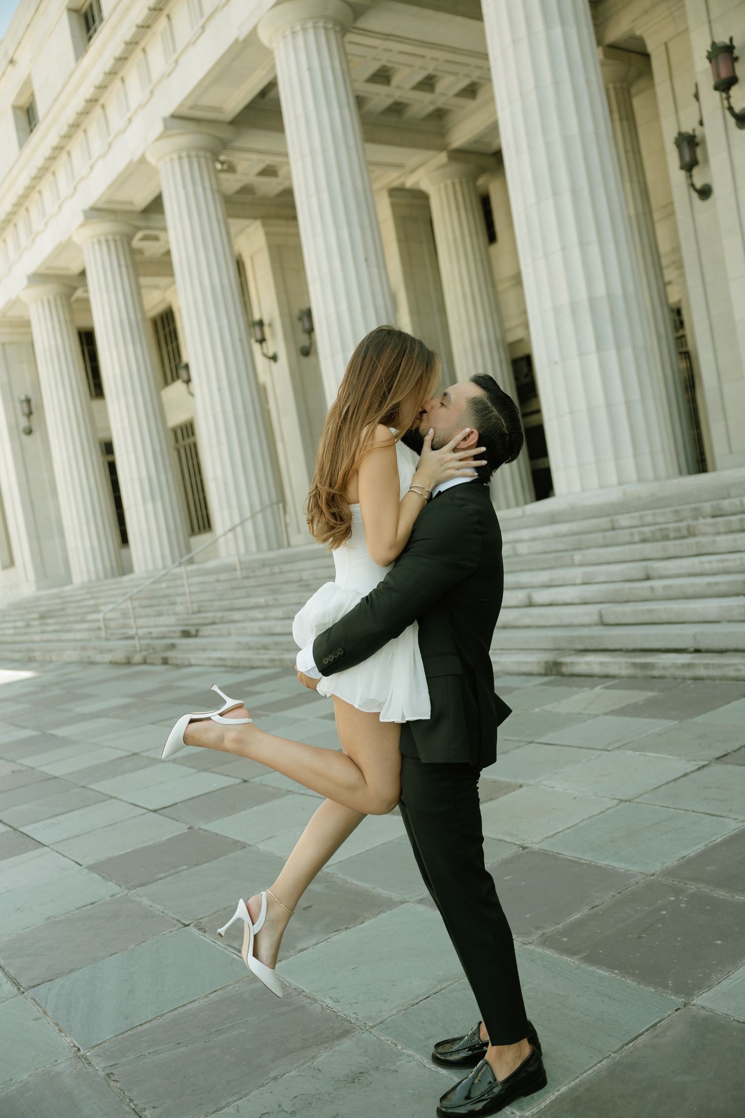 Romantic couple sharing a kiss on steps of neoclassical building, bride in white mini dress lifted by groom in tuxedo.