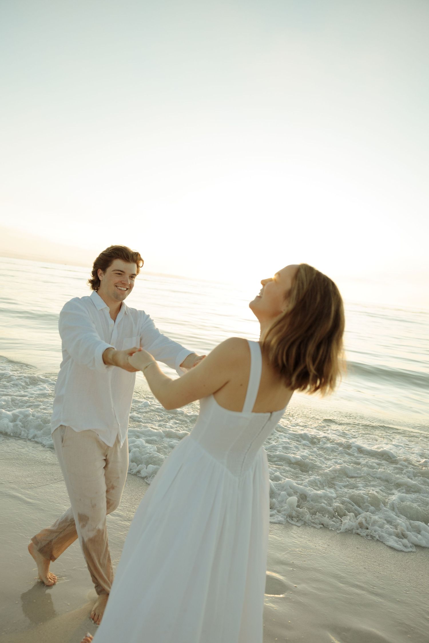 Couple in white clothing dancing joyfully on a sunset beach with ocean waves in the background.