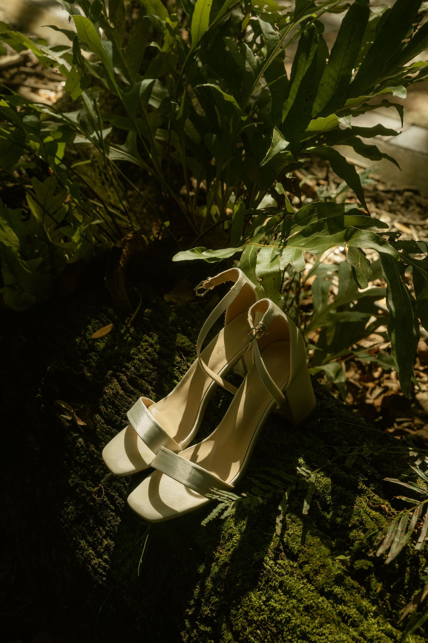 Elegant cream bridal heeled sandals resting on moss-covered ground surrounded by lush green tropical foliage.