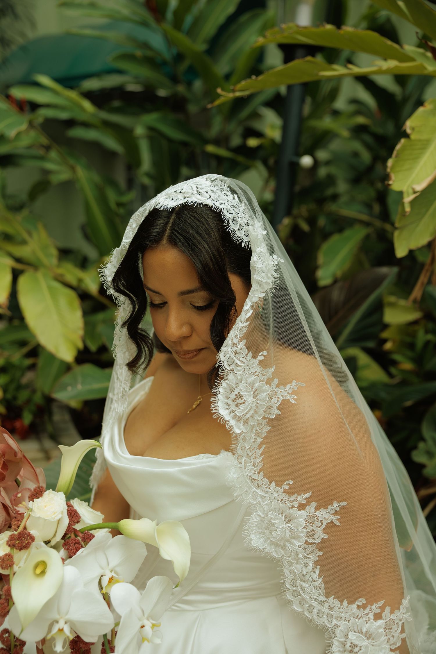 Bride in white lace veil and gown holding white orchid bouquet, posing outdoors among lush green tropical plants.