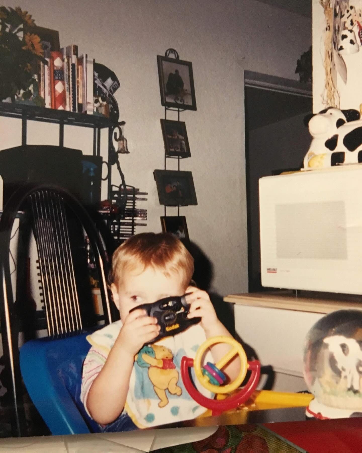 A young child in a blue chair holds a retro camera up to their face with a pet rabbit visible nearby.