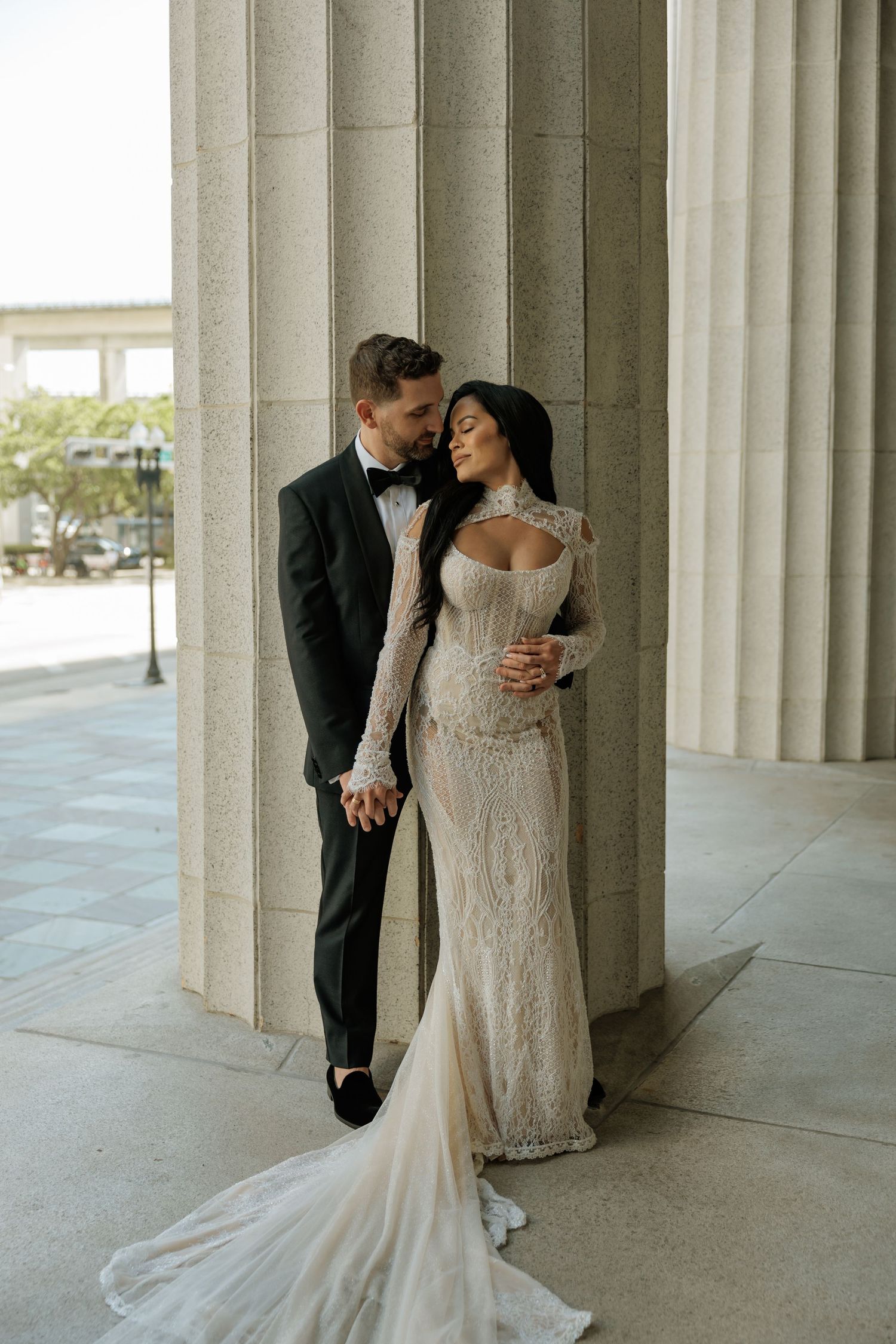 Romantic wedding couple embraces between elegant white stone columns in an outdoor colonnade.