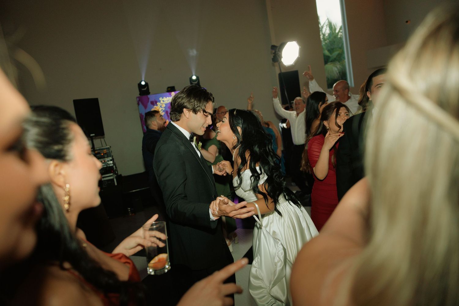 Couple sharing a kiss on a crowded wedding reception dance floor surrounded by guests in formal attire.