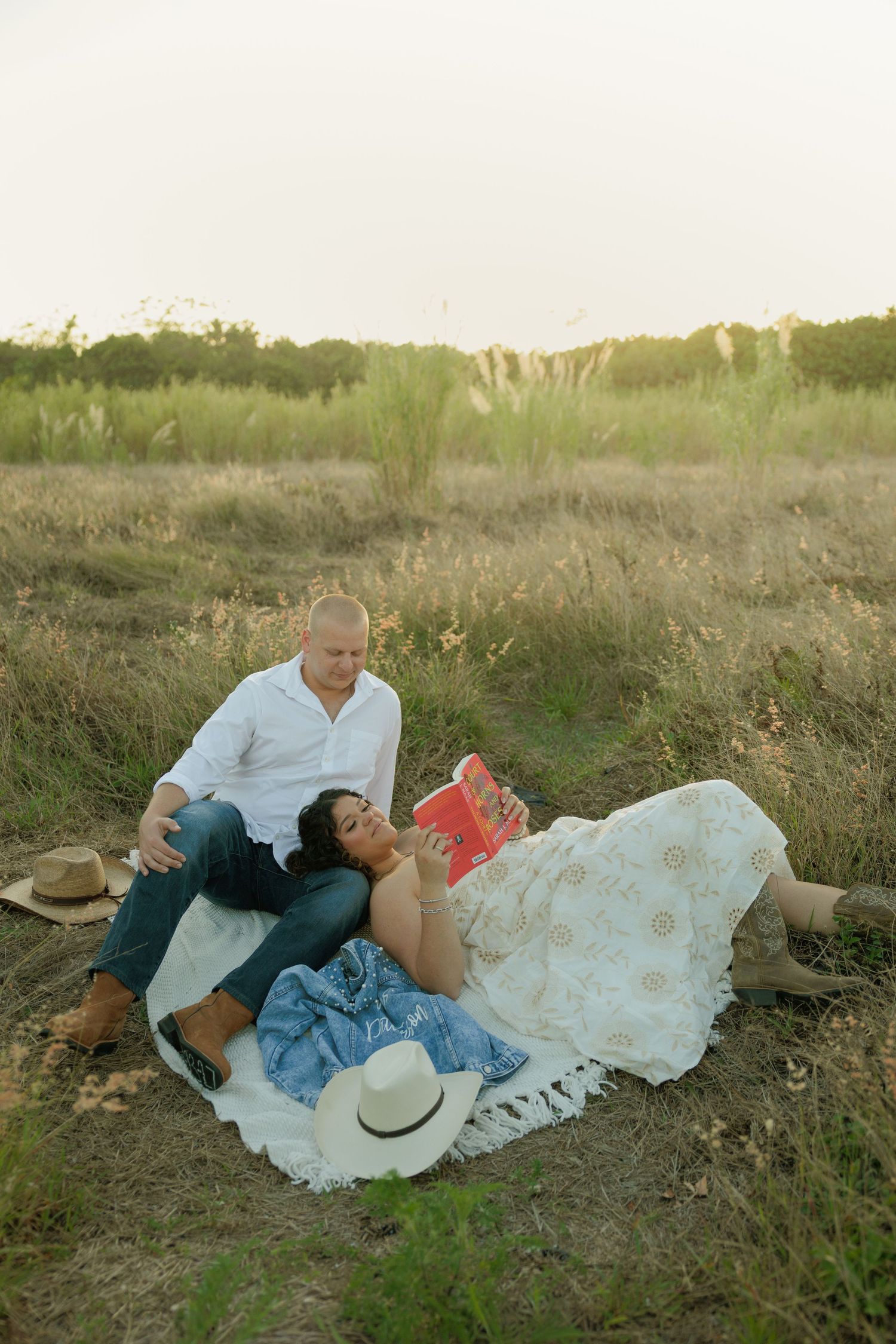 Couple relaxing in a grassy field at sunset, woman reading a red book while resting on man's lap on a blue blanket.