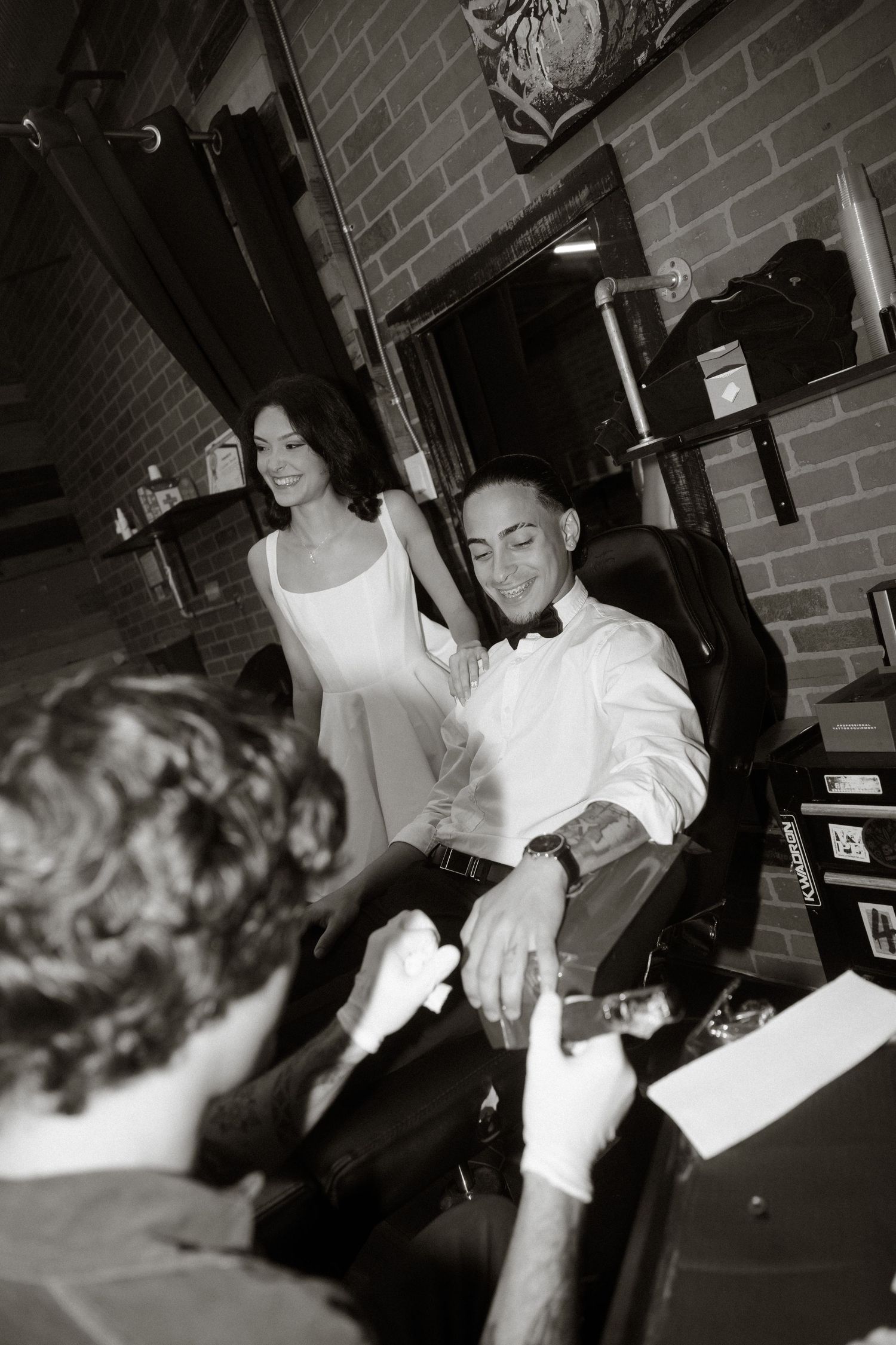 Black and white photo shows people in formal wear at a bar with exposed brick walls and shelving behind them.