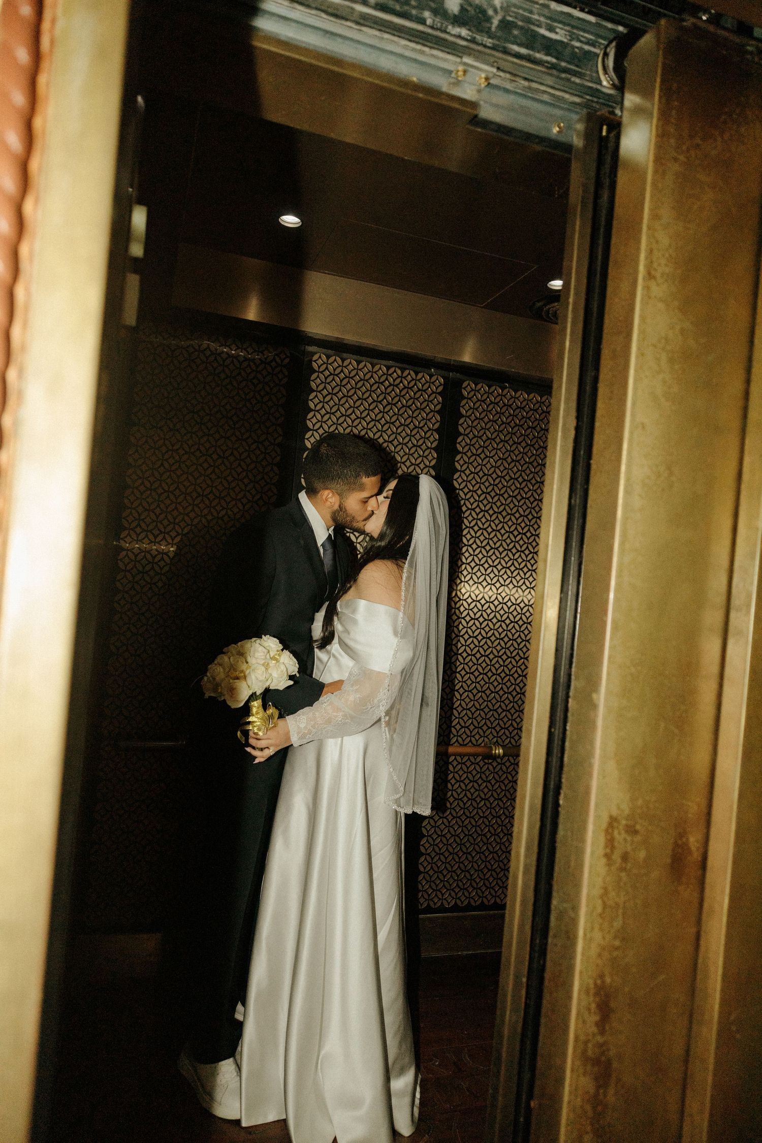 A couple shares an intimate embrace in an elevator, with the bride wearing a white gown and holding a bouquet.