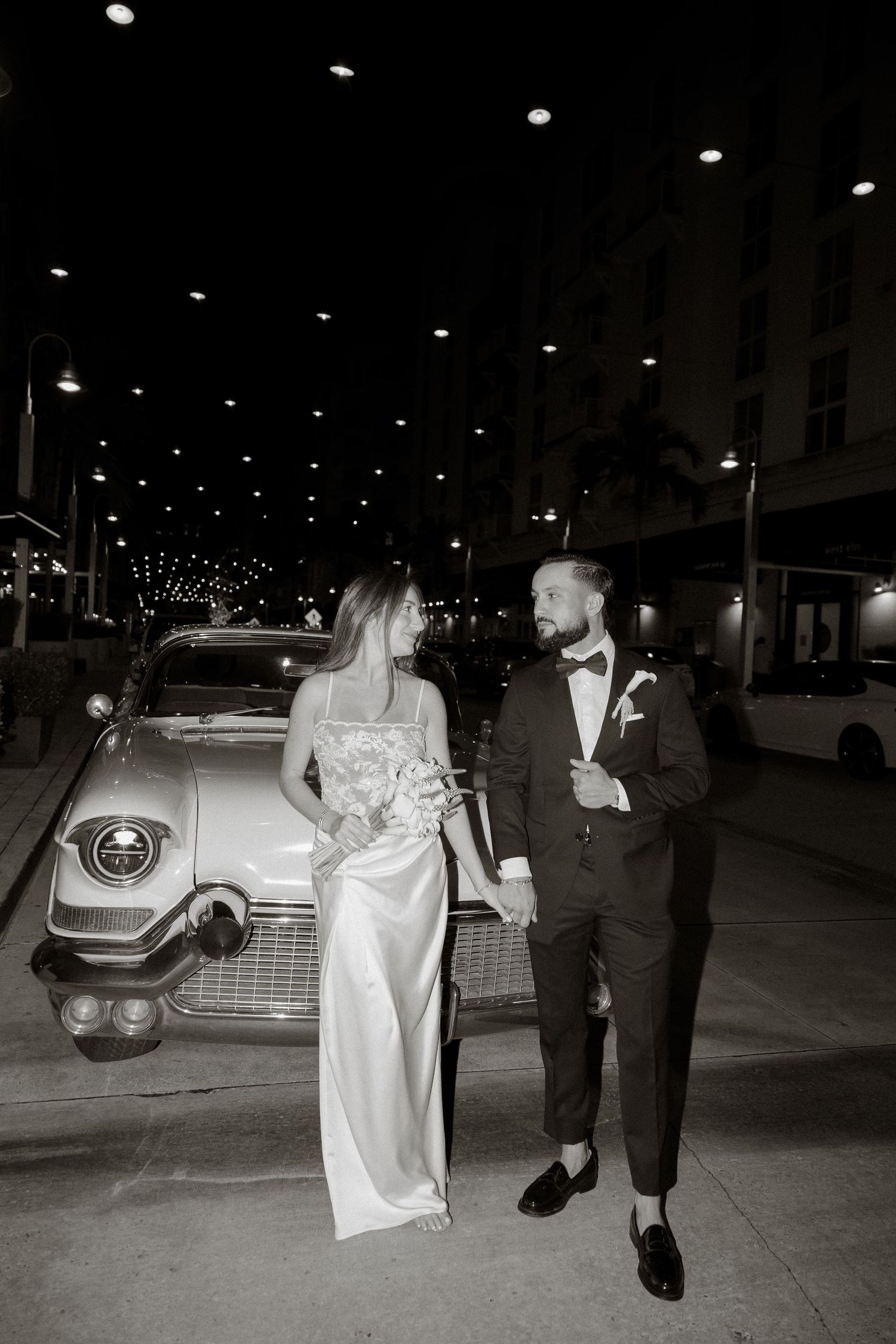 Black and white photo of a bride and groom posing beside a vintage car on a city street at night.