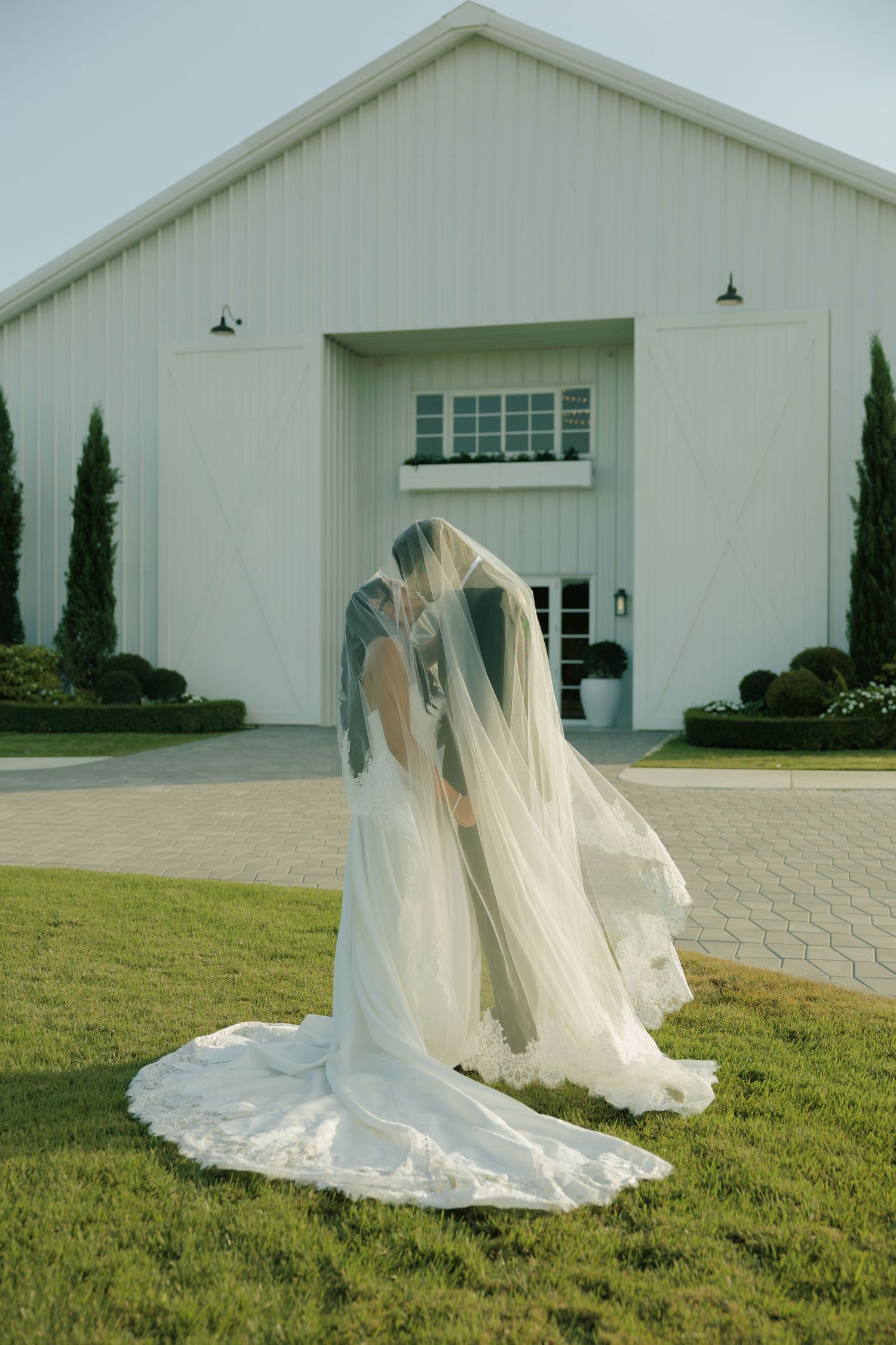 Two brides sharing a kiss under a flowing veil in front of a white barn wedding venue.