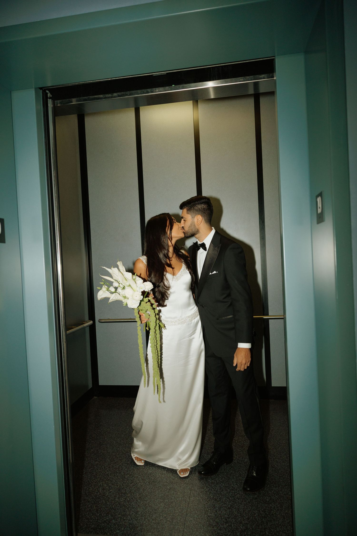 Bride and groom sharing a kiss in an elevator, bride holding white floral bouquet, groom in black tuxedo.