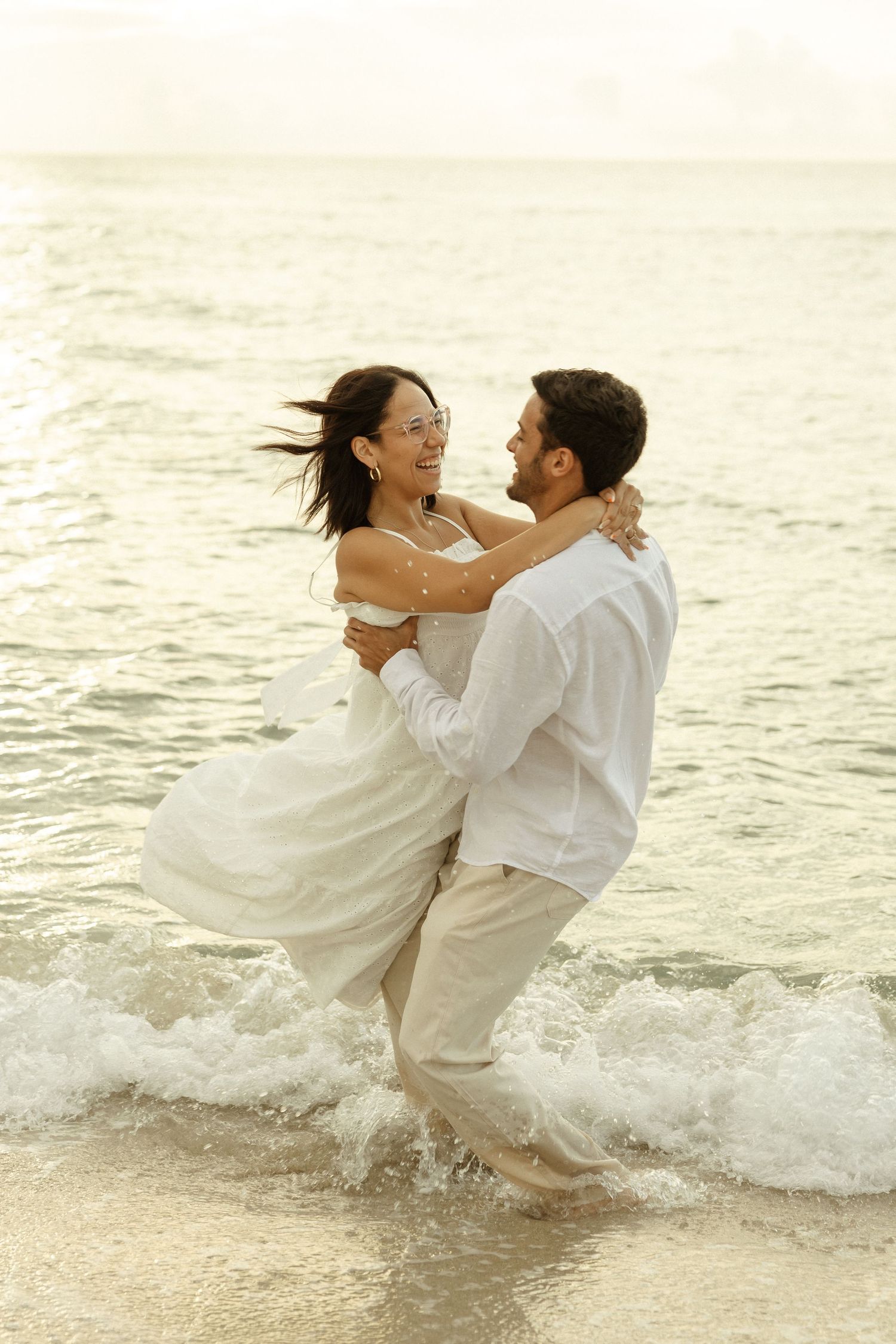 Romantic couple dressed in white embraces while standing in ocean waves during sunset at the beach.