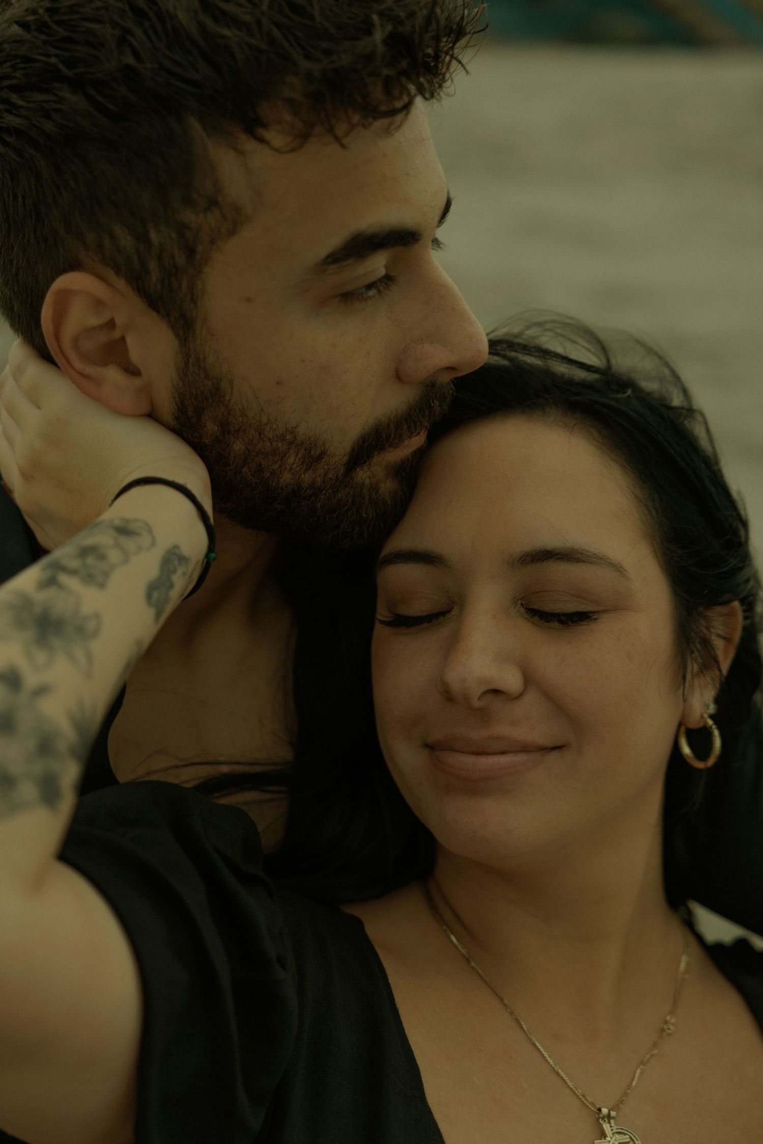 A tattooed man tenderly embraces a smiling dark-haired woman wearing gold hoop earrings and a necklace.
